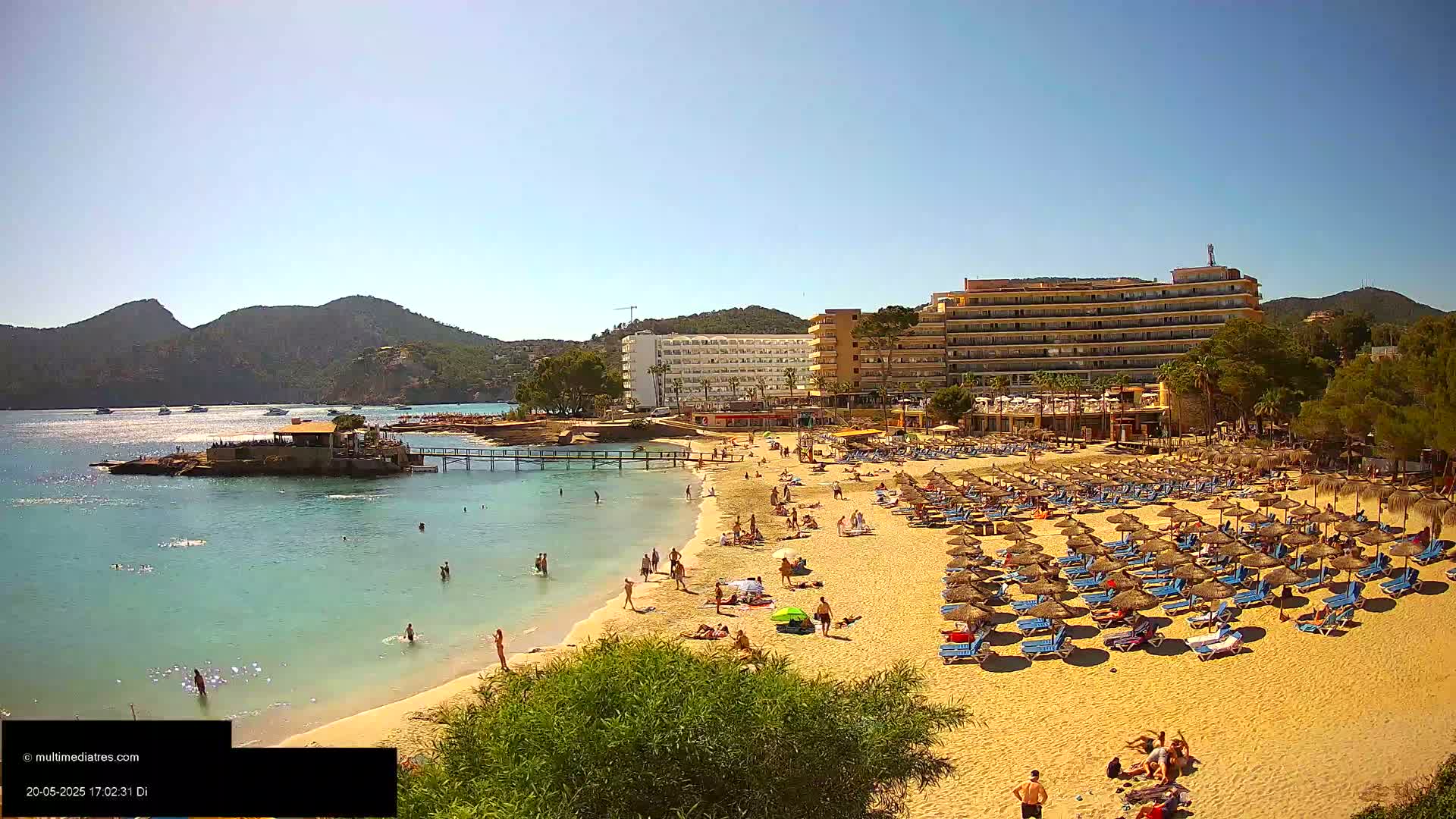 A sandy beach with numerous umbrellas and sunbathers is situated in front of a large hotel, overlooking calm, clear water and mountains under a sunny sky.
