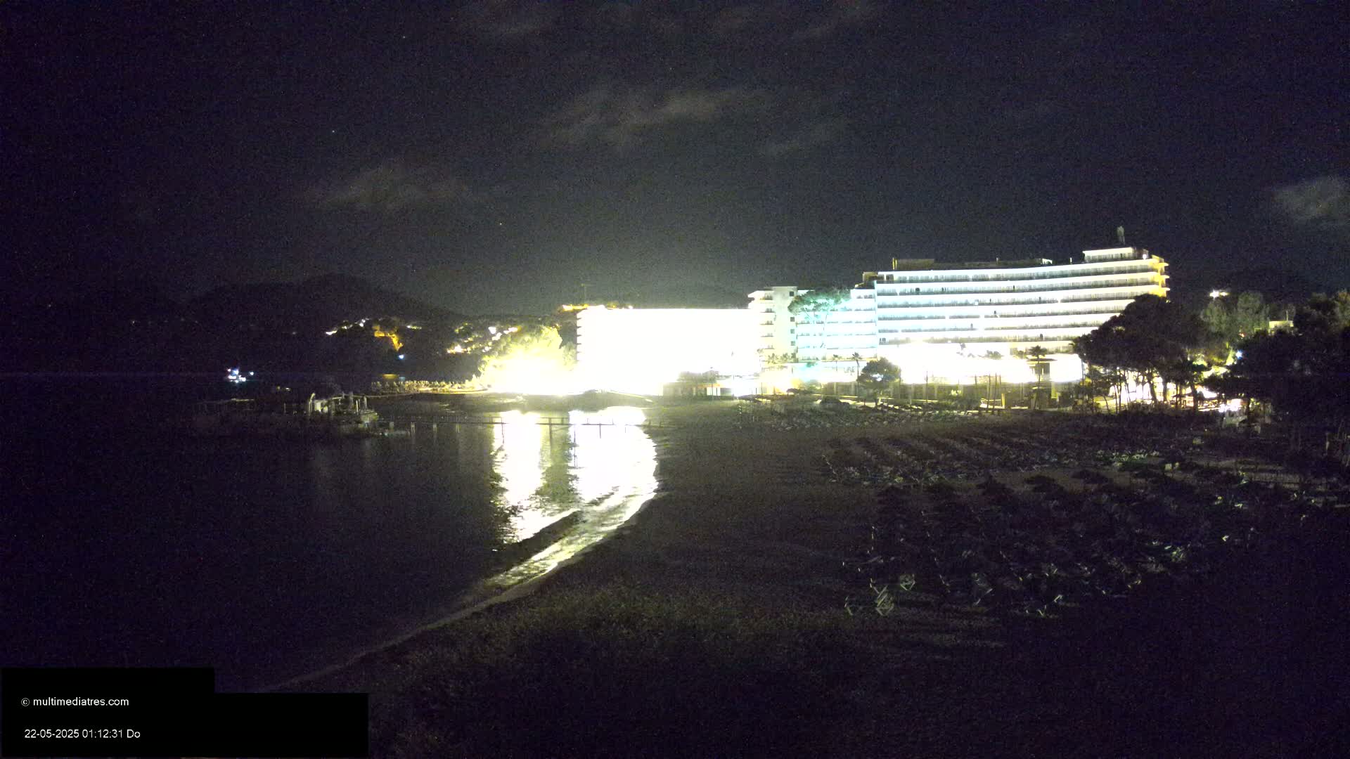 A brightly lit hotel overlooks a dark beach at night, with calm water reflecting the lights.