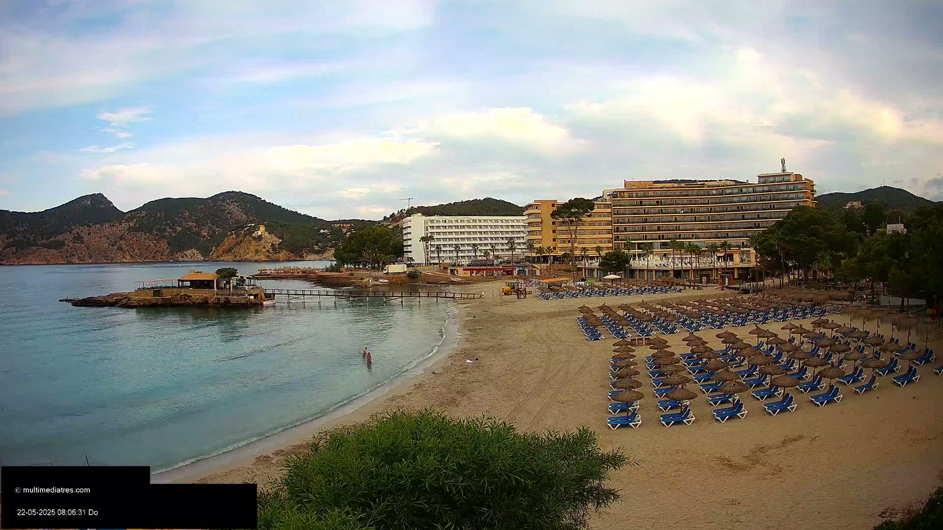 A sandy beach with numerous rows of thatched umbrellas and lounge chairs sits below a pair of large hotels and overlooks a calm bay surrounded by hills under a partly cloudy sky.