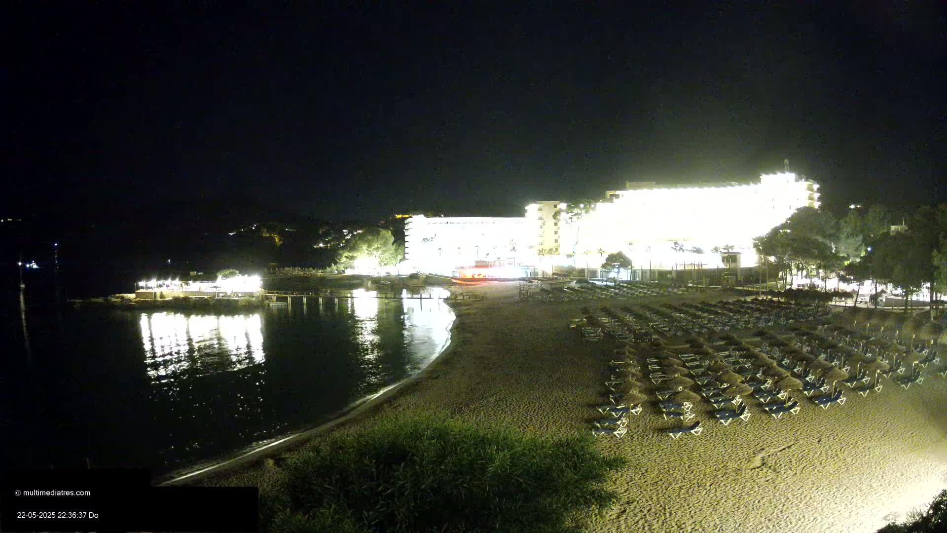 A brightly lit hotel overlooks a sandy beach at night, with rows of lounge chairs and calm water reflecting the lights.