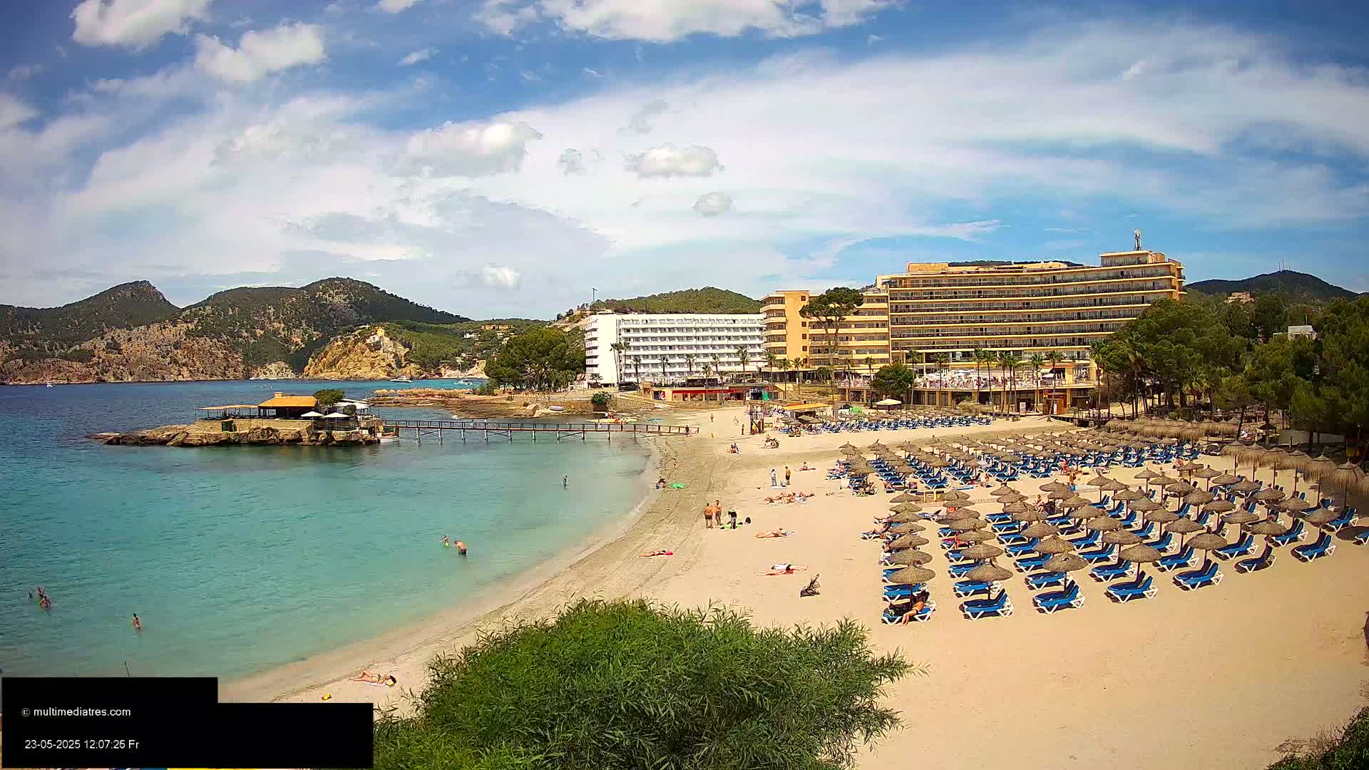 A sandy beach with numerous umbrellas and lounge chairs, bordered by a turquoise ocean,  a small island with a building, and hotels backed by hills under a partly cloudy blue sky.