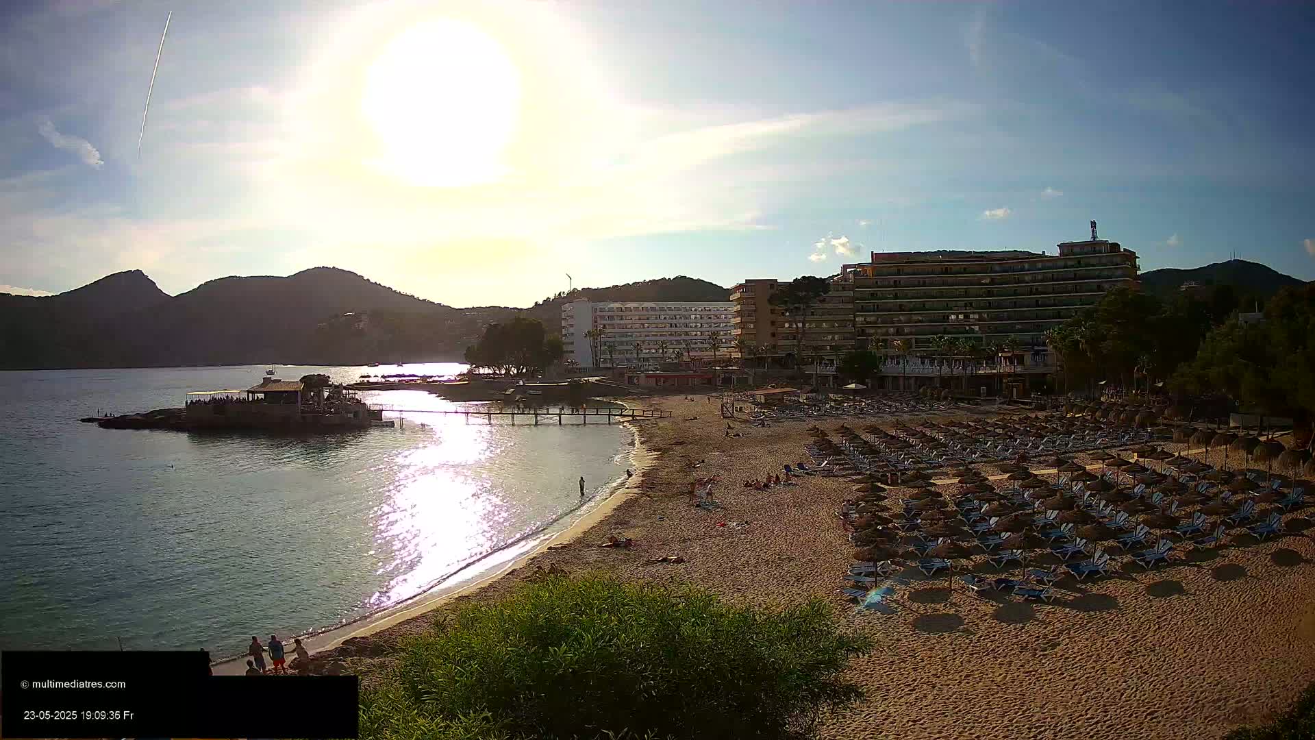 A sandy beach with numerous lounge chairs and umbrellas sits below a large hotel, all overlooking calm water with mountains in the background under a bright, sunny sky.