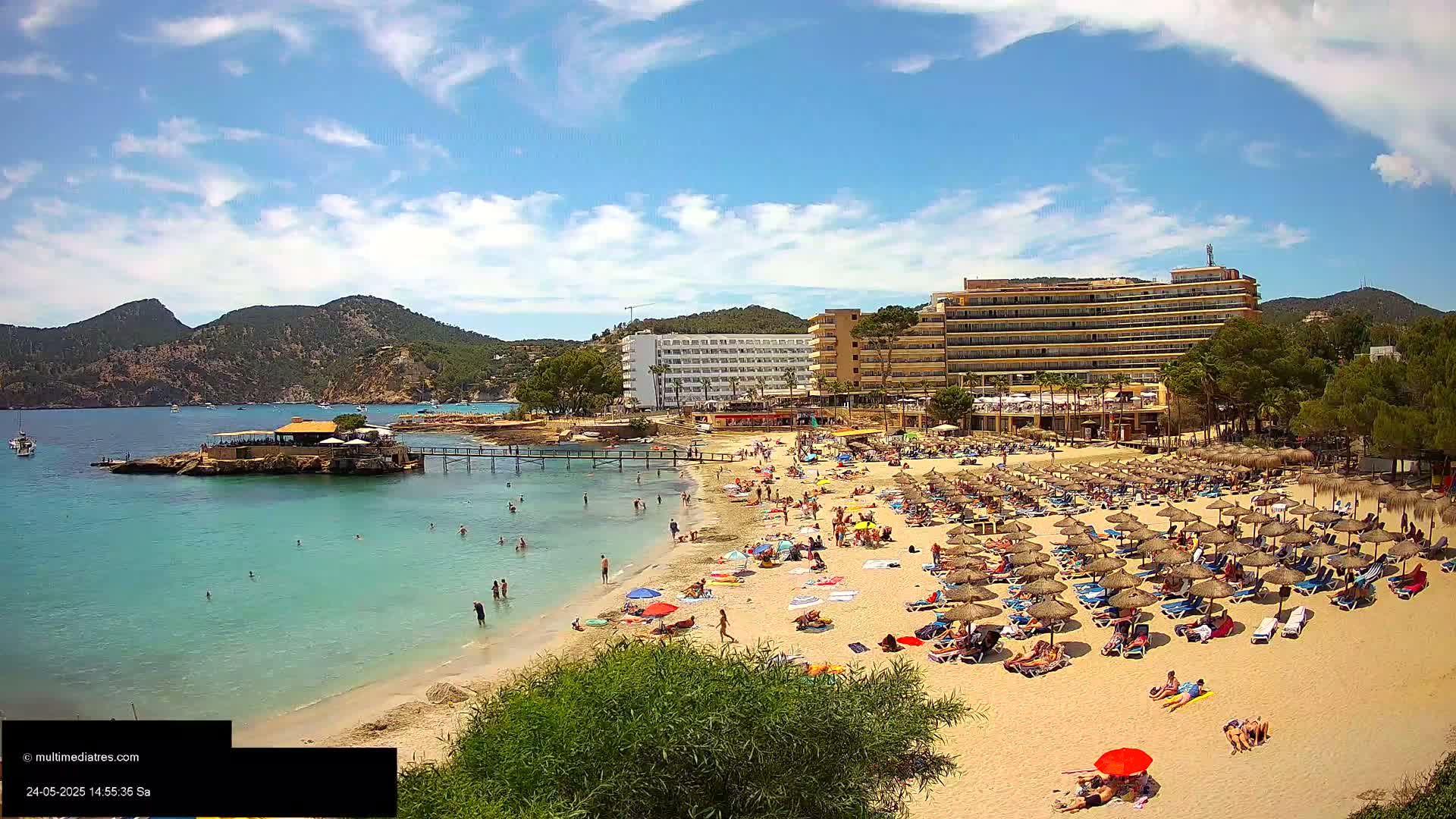 A sandy beach with many people sunbathing under thatched umbrellas, a calm turquoise sea, a small restaurant on an islet, and large hotels behind it, all under a sunny sky with some clouds.