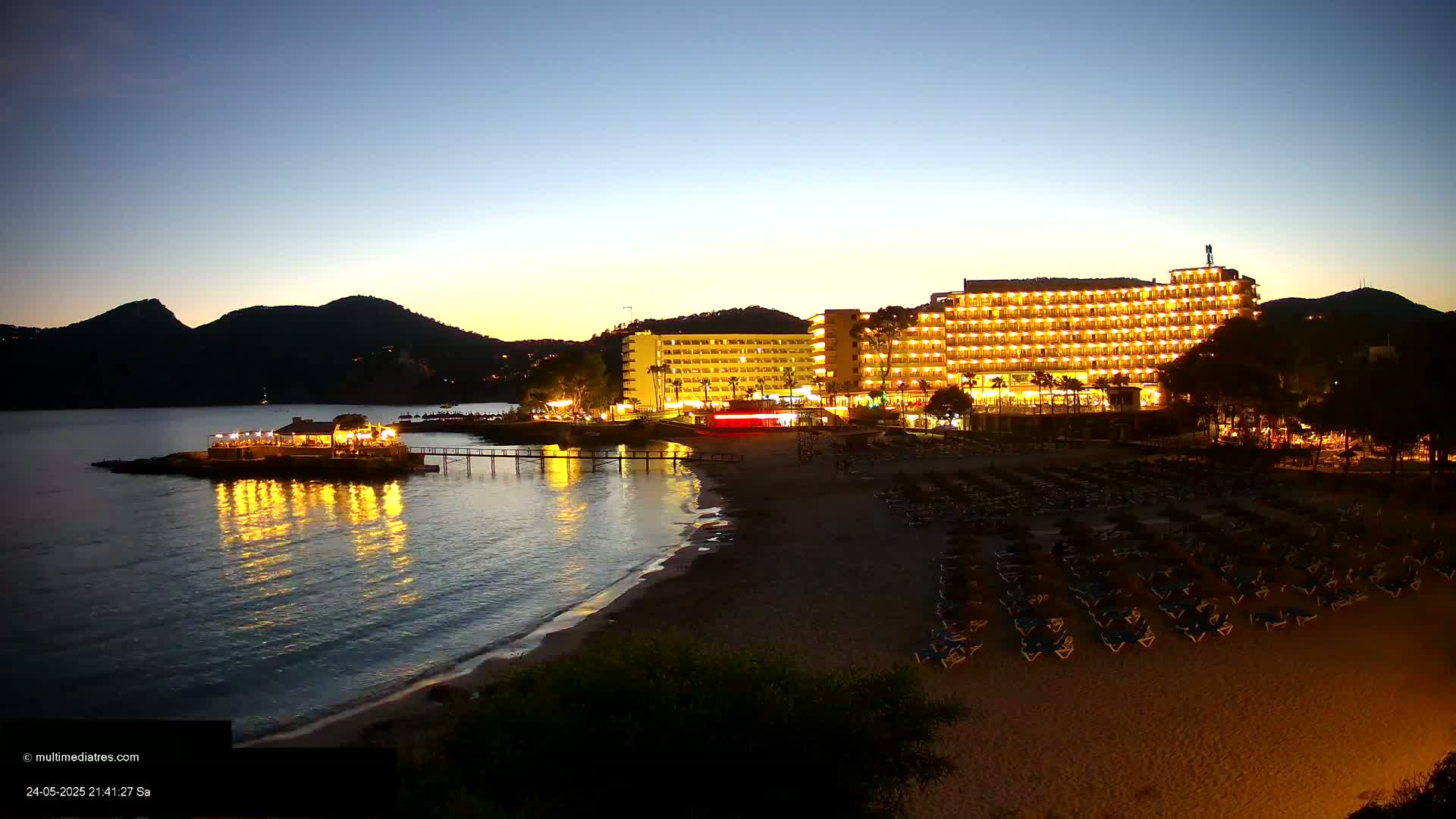 A brightly lit hotel stands on a sandy beach at dusk, next to a calm body of water reflecting the lights, with a smaller illuminated building on a pier in the water.