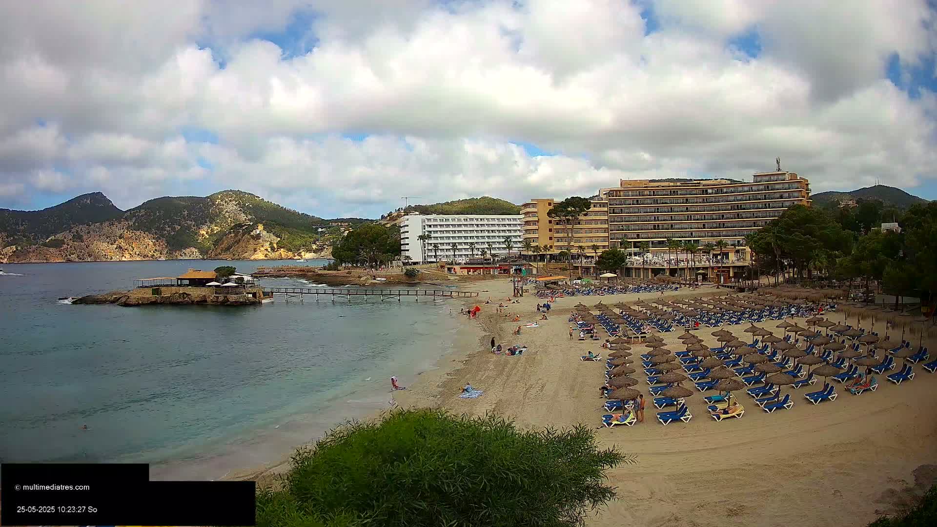 A sandy beach with numerous blue lounge chairs and thatched umbrellas sits in front of several hotels, a small island with a building, and mountains under a partly cloudy sky.