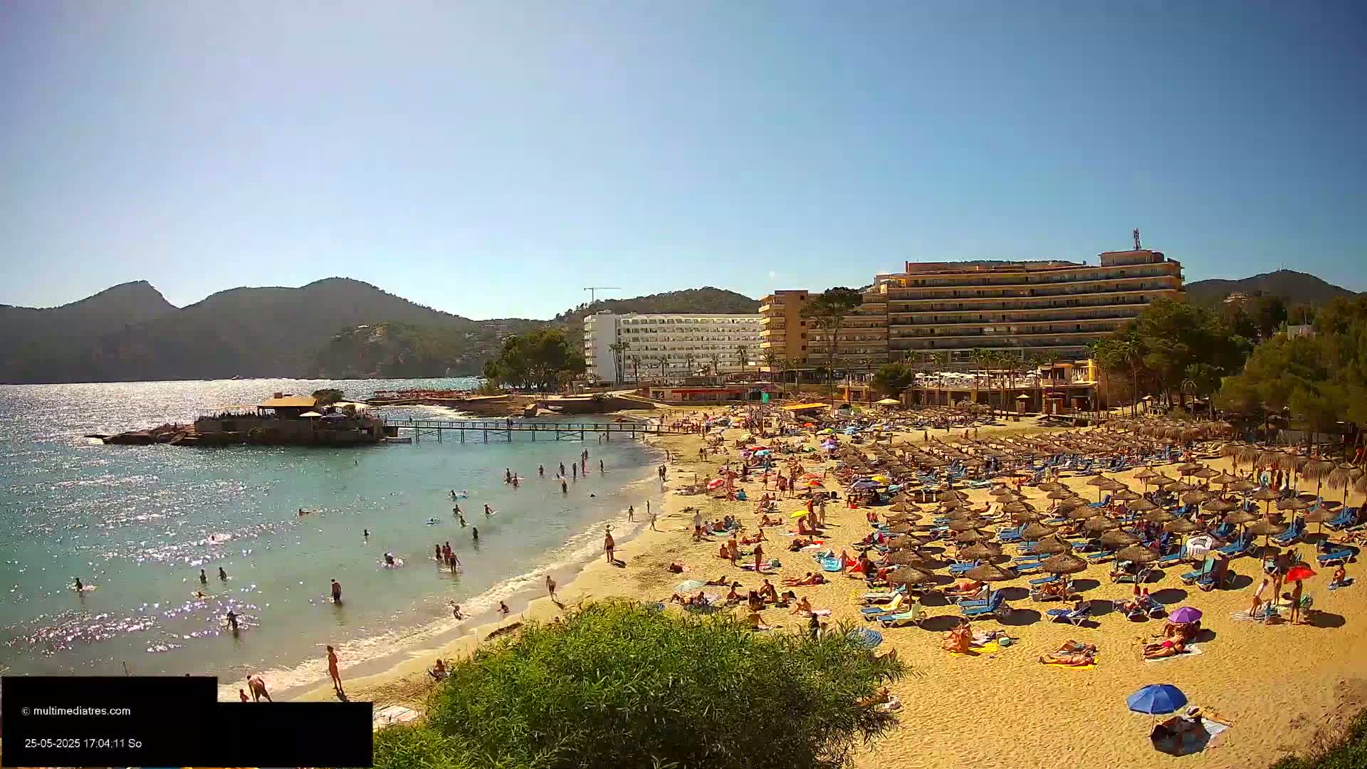 A sunny beach scene shows a crowded sandy beach with numerous umbrellas and beachgoers, a calm sea with swimmers, and a hotel and other buildings behind it, all backed by mountains under a clear blue sky.