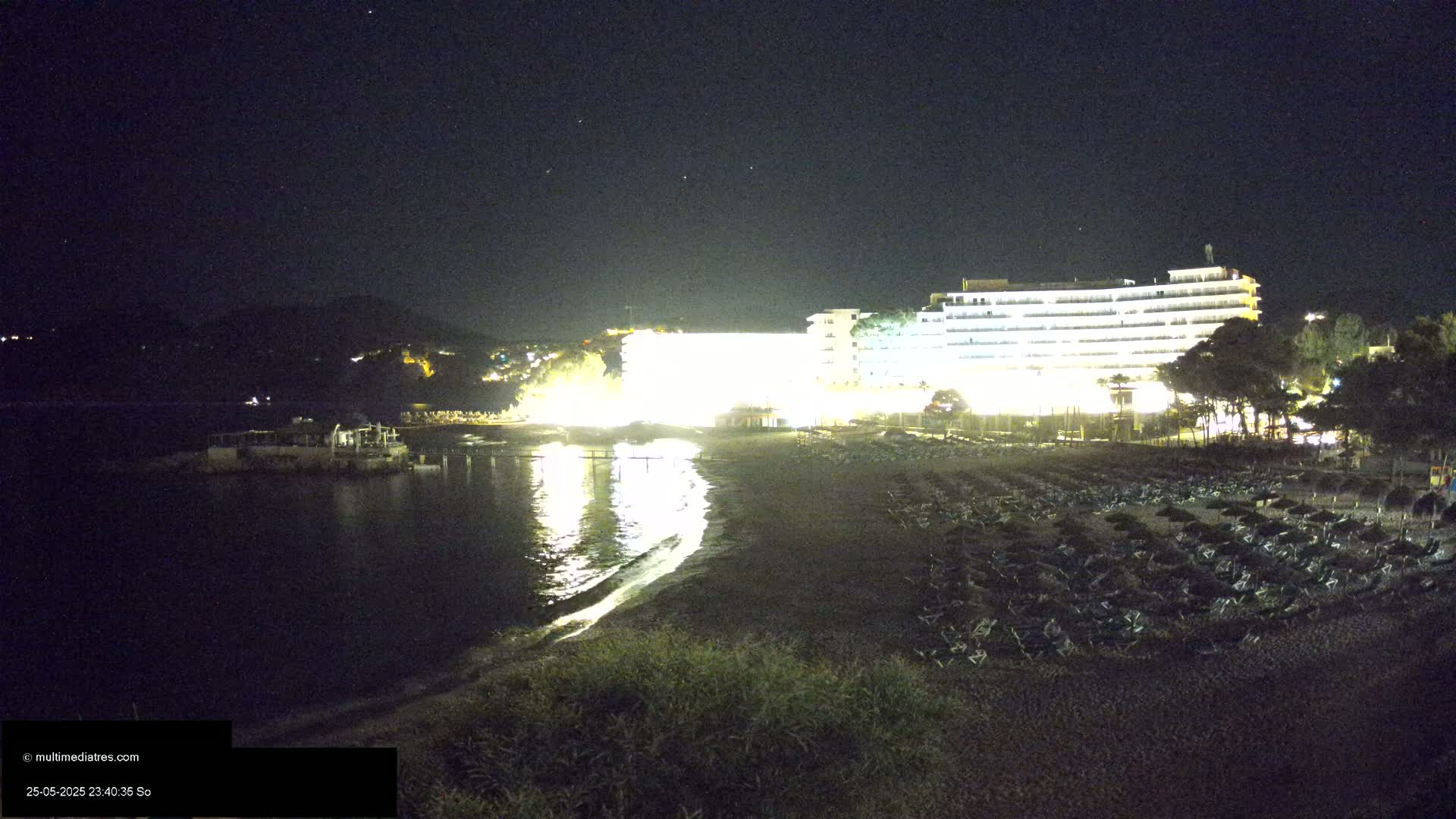 A brightly lit hotel overlooks a dark beach at night, with numerous lounge chairs and umbrellas set up on the sand, under a clear, starry sky.