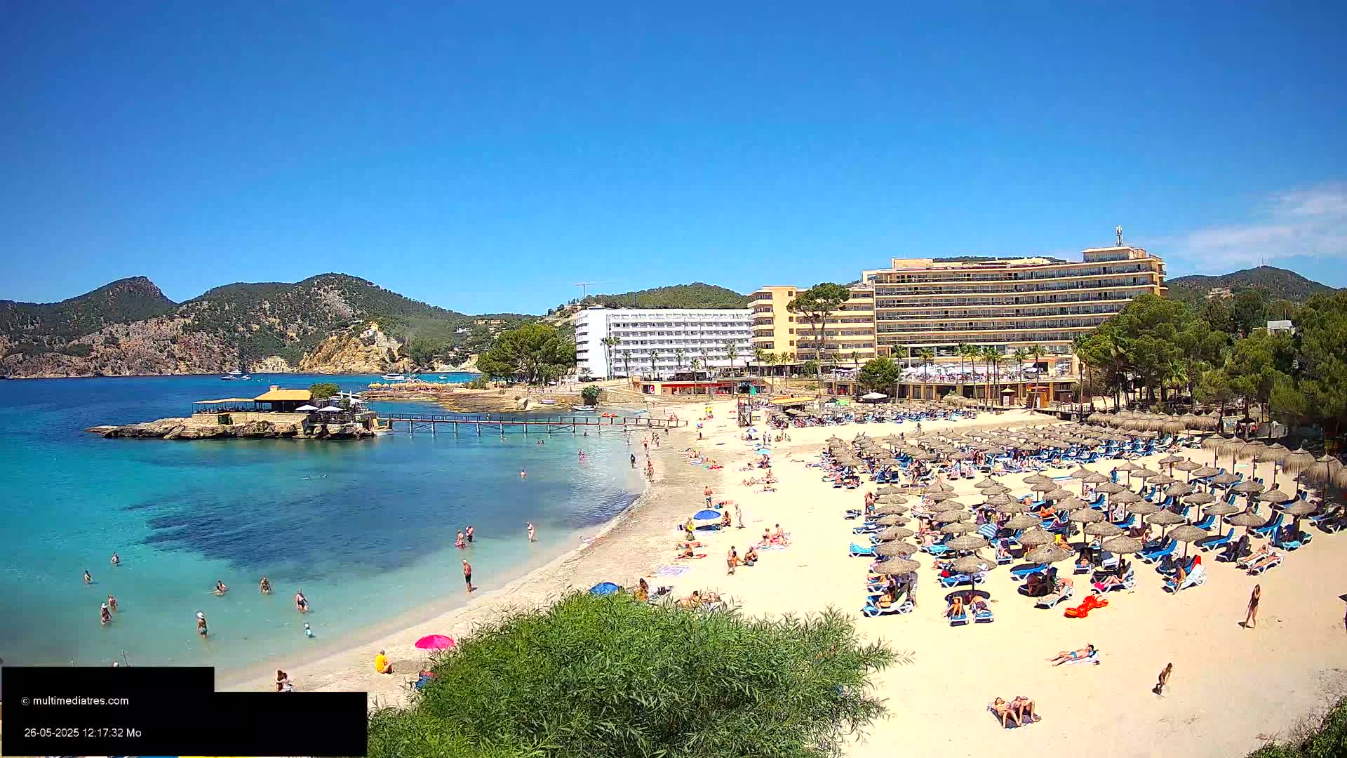 A sandy beach with numerous umbrellas and sunbathers is situated next to a calm, clear turquoise sea, backed by hotels and green hills under a bright sunny sky.