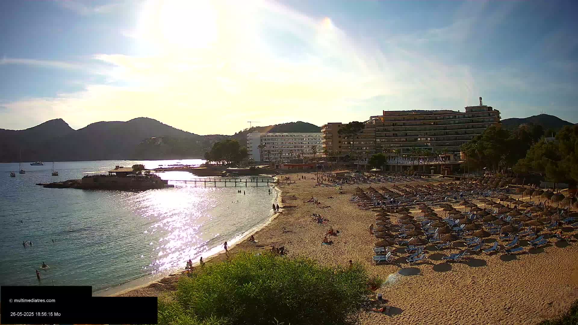 A sunny beach scene shows a sandy shore lined with numerous beach umbrellas and lounge chairs, backed by hotels and a calm bay with boats visible in the distance, all nestled against a mountainous backdrop.