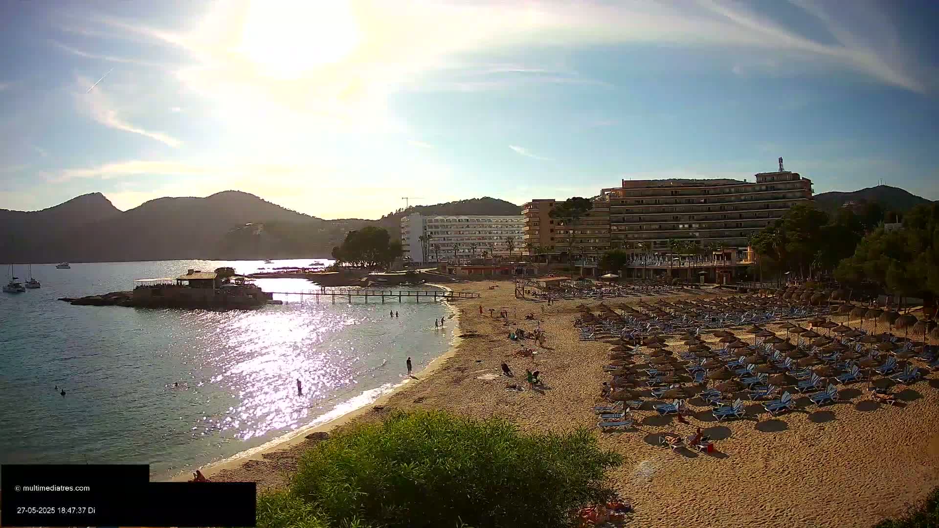 A sunny beach scene shows a sandy shoreline lined with numerous beach umbrellas and lounge chairs, adjacent to a calm bay with a small pier and several buildings, backed by hills under a mostly clear sky.