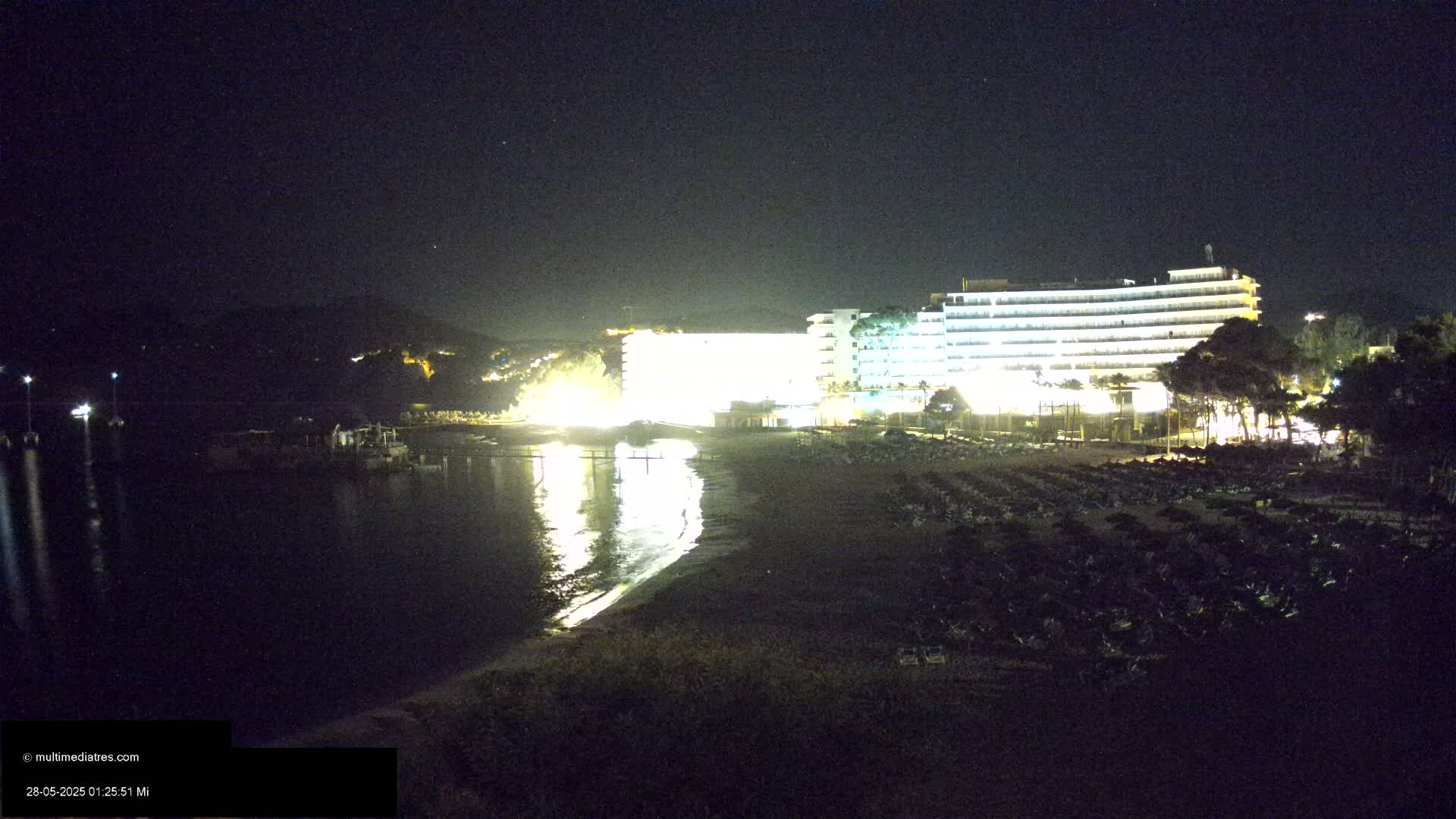 A brightly lit hotel overlooks a dark sandy beach and calm, reflective water at night.