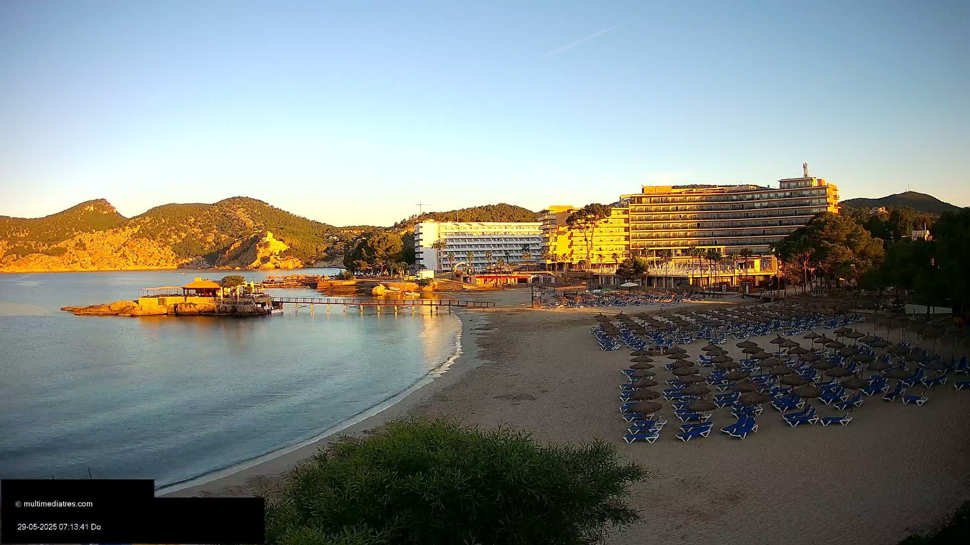 A sandy beach with numerous lounge chairs and umbrellas sits in front of a large hotel complex,  all under a clear, sunrise sky, with calm water and hills in the background.
