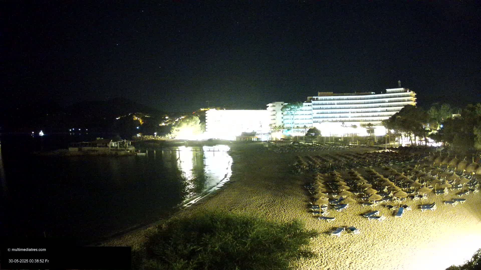 A sandy beach at night, lined with rows of beach umbrellas and chairs, next to a brightly lit hotel, with calm water reflecting the lights in the background under a clear, starry sky.