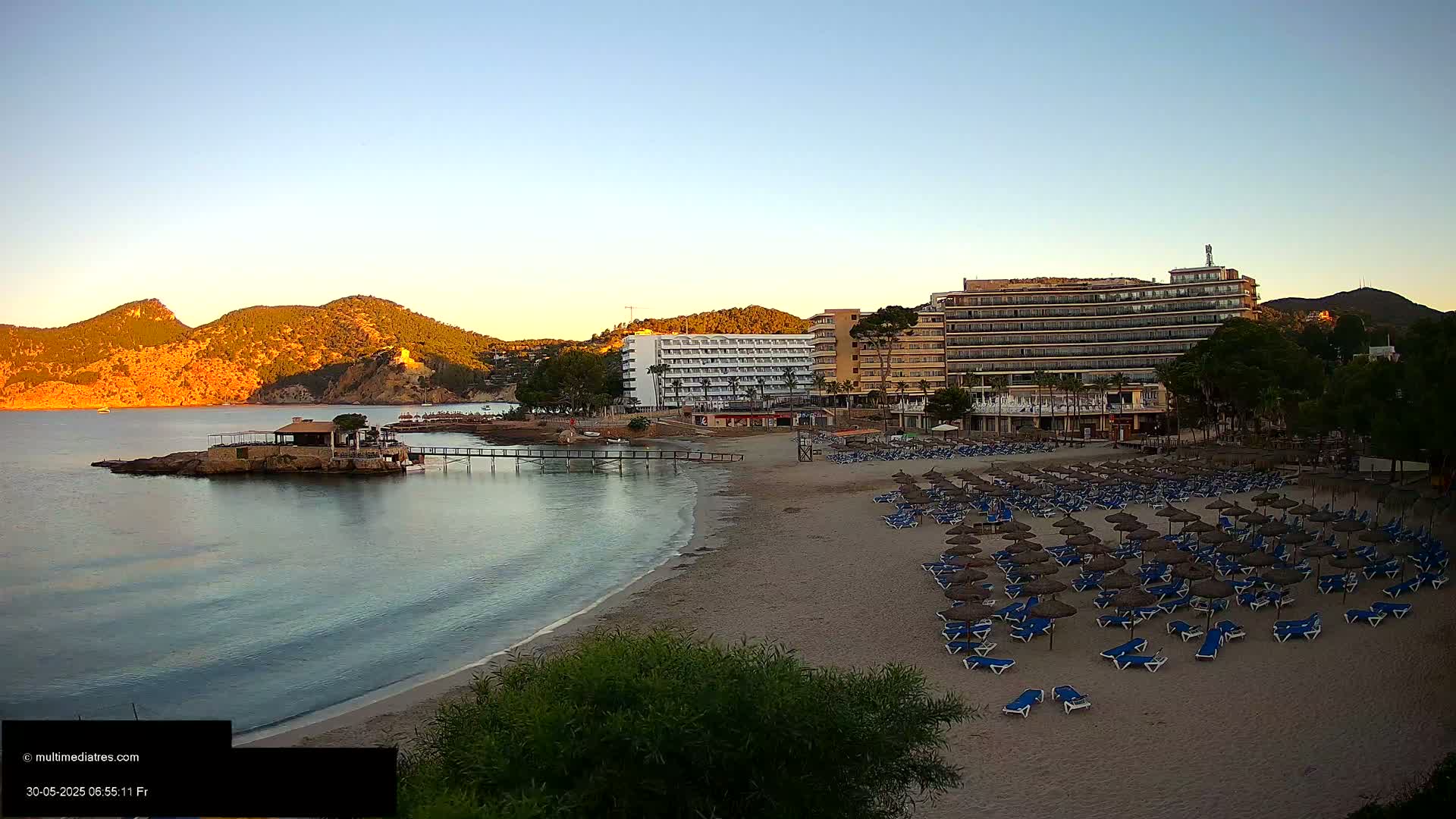 A sandy beach with numerous unoccupied beach umbrellas and lounge chairs sits in front of several hotels,  with calm, clear water lapping the shore under a clear, sunrise sky and hills in the background.