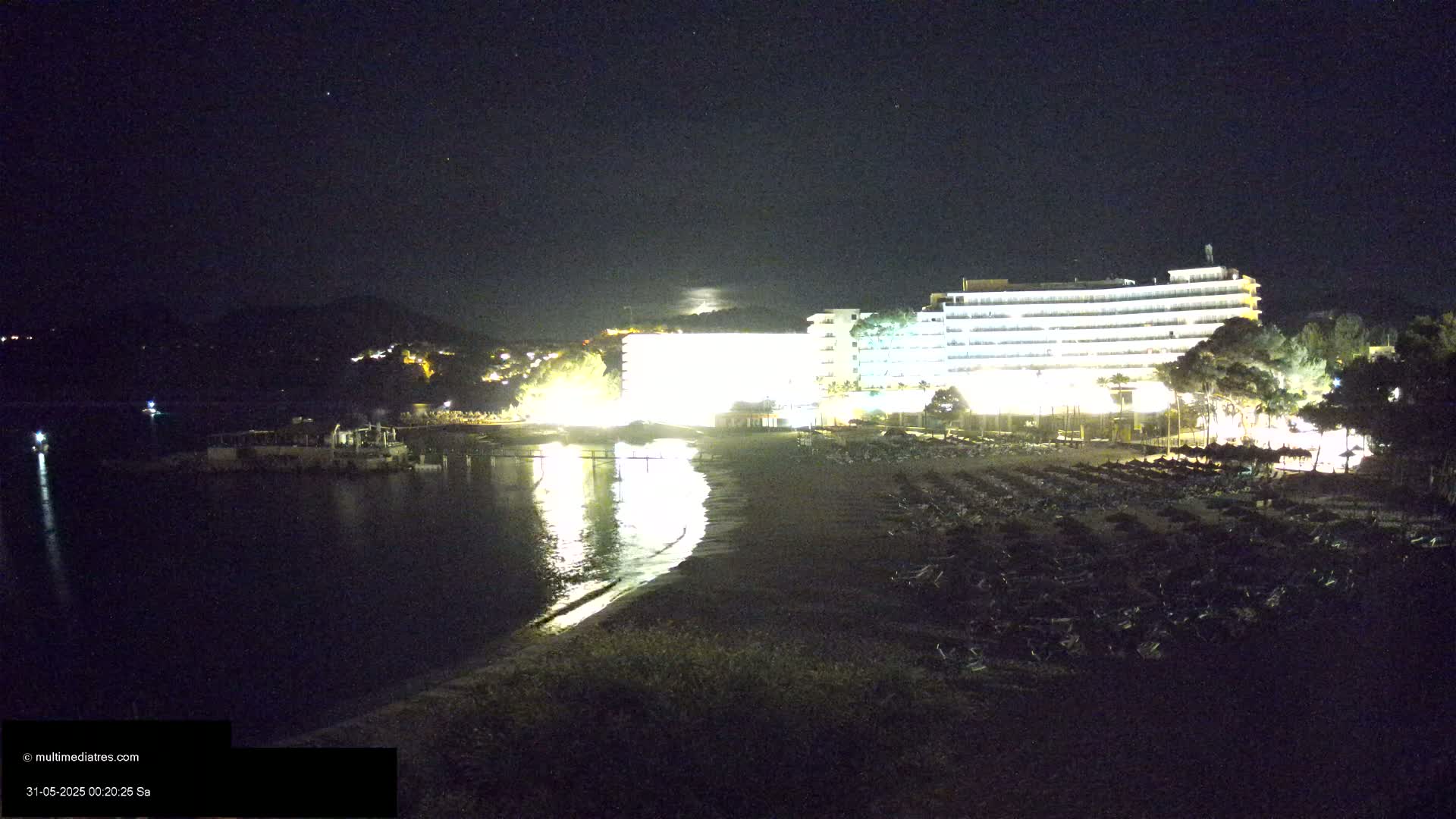 A brightly lit hotel stands on a dark beach at night next to calm water under a clear, starry sky.