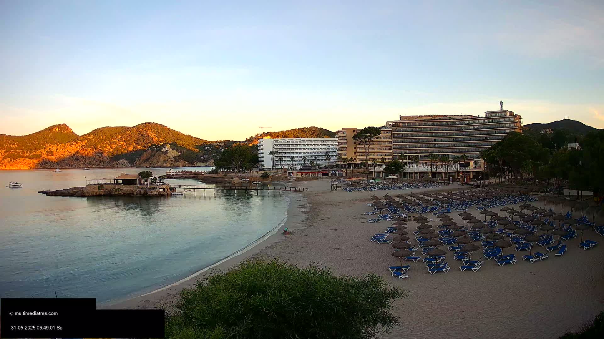 A sandy beach with numerous empty lounge chairs and umbrellas sits in front of several hotels, overlooking a calm bay and golden-hued hills under a clear, sunrise sky.