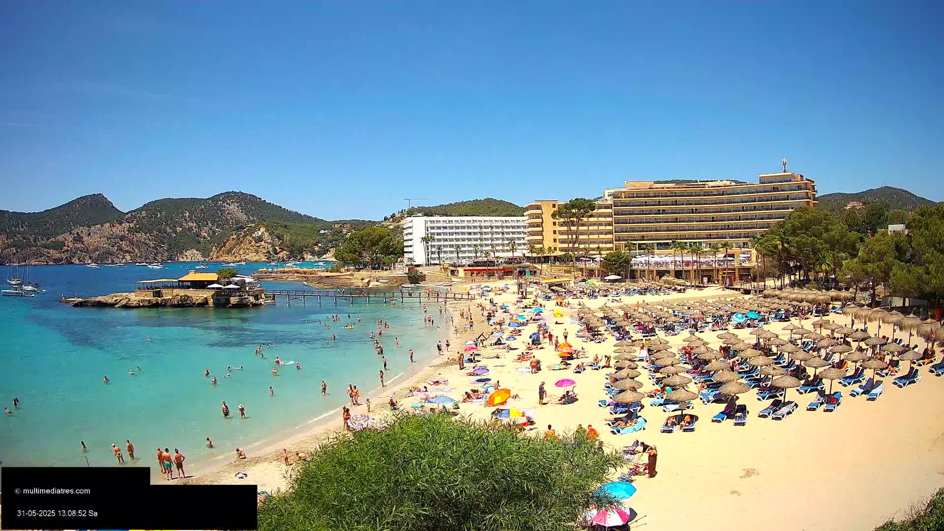 A sunny beach scene shows numerous beachgoers relaxing under umbrellas near a hotel and turquoise water, with a small island and mountains in the background.