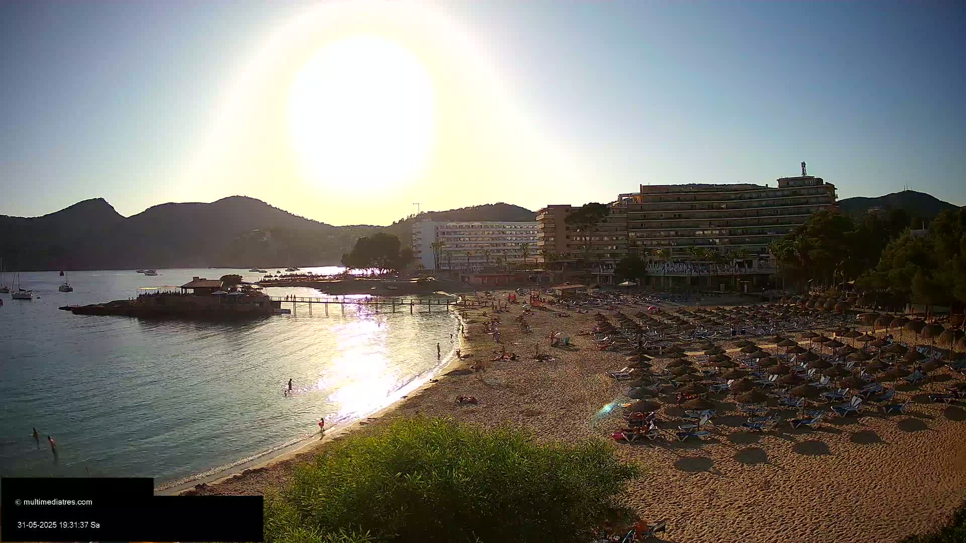 A sandy beach with numerous umbrellas and lounge chairs is situated next to a calm bay, under a bright, sunny sky, with hotels and mountains in the background.
