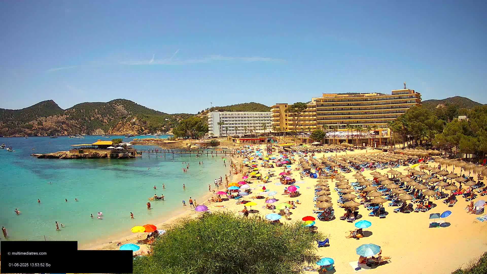 A sunny beach scene shows a crowded sandy beach with numerous colorful umbrellas and lounge chairs, turquoise water, and a large hotel in the background.