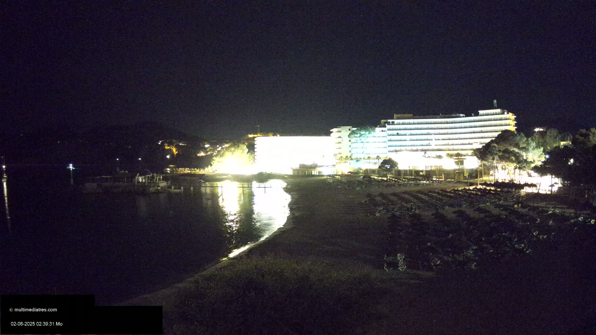 A brightly lit hotel stands on a dark beach at night next to a calm body of water.