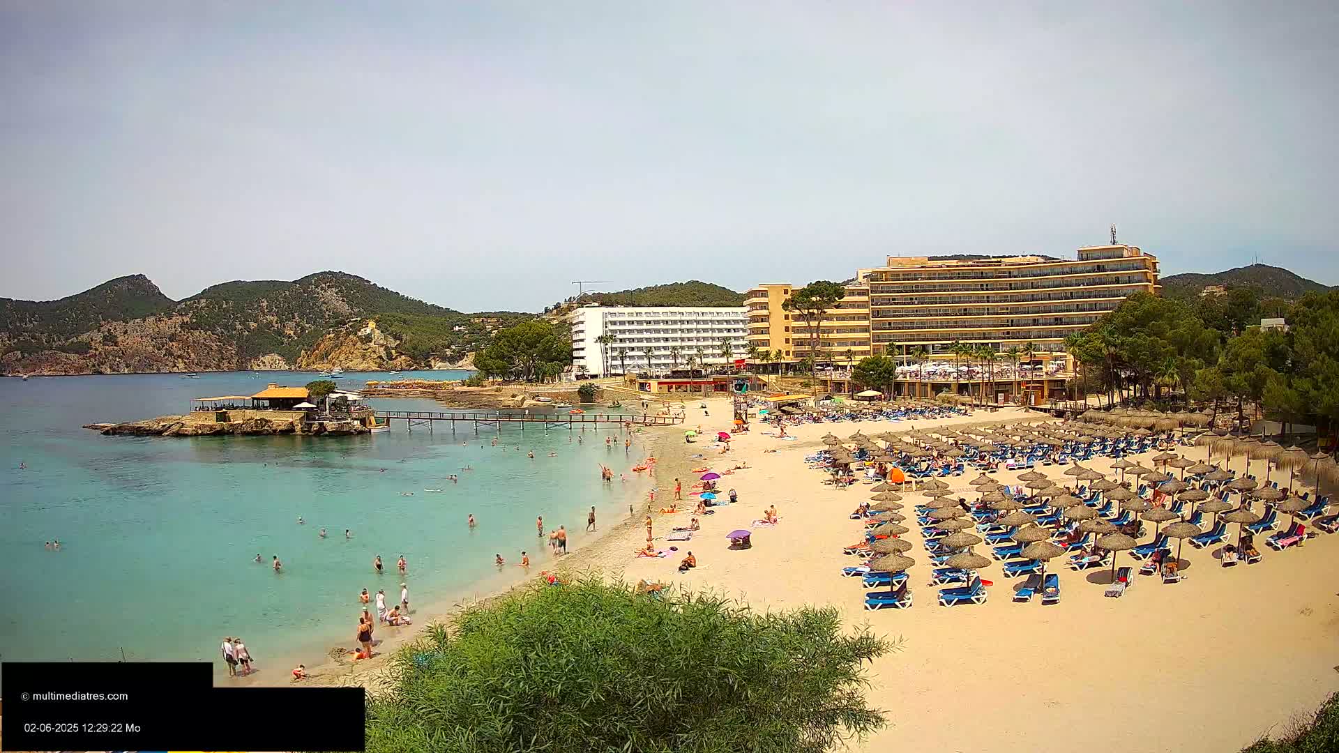 A sandy beach with numerous umbrellas and lounge chairs, many people swimming and sunbathing, overlooks a calm, turquoise bay with a small restaurant on an islet and large hotels behind it, under a bright, sunny sky.
