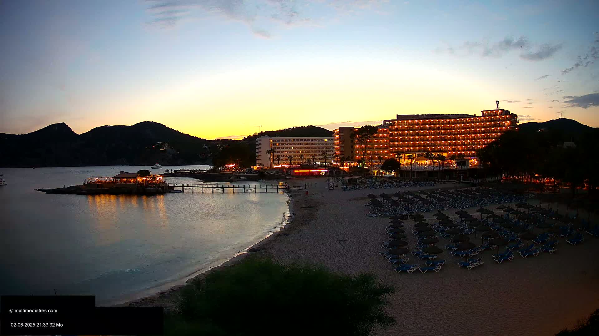 A beach at dusk with a lit-up hotel and beach umbrellas, a small restaurant on a pier, and dark mountains in the background.