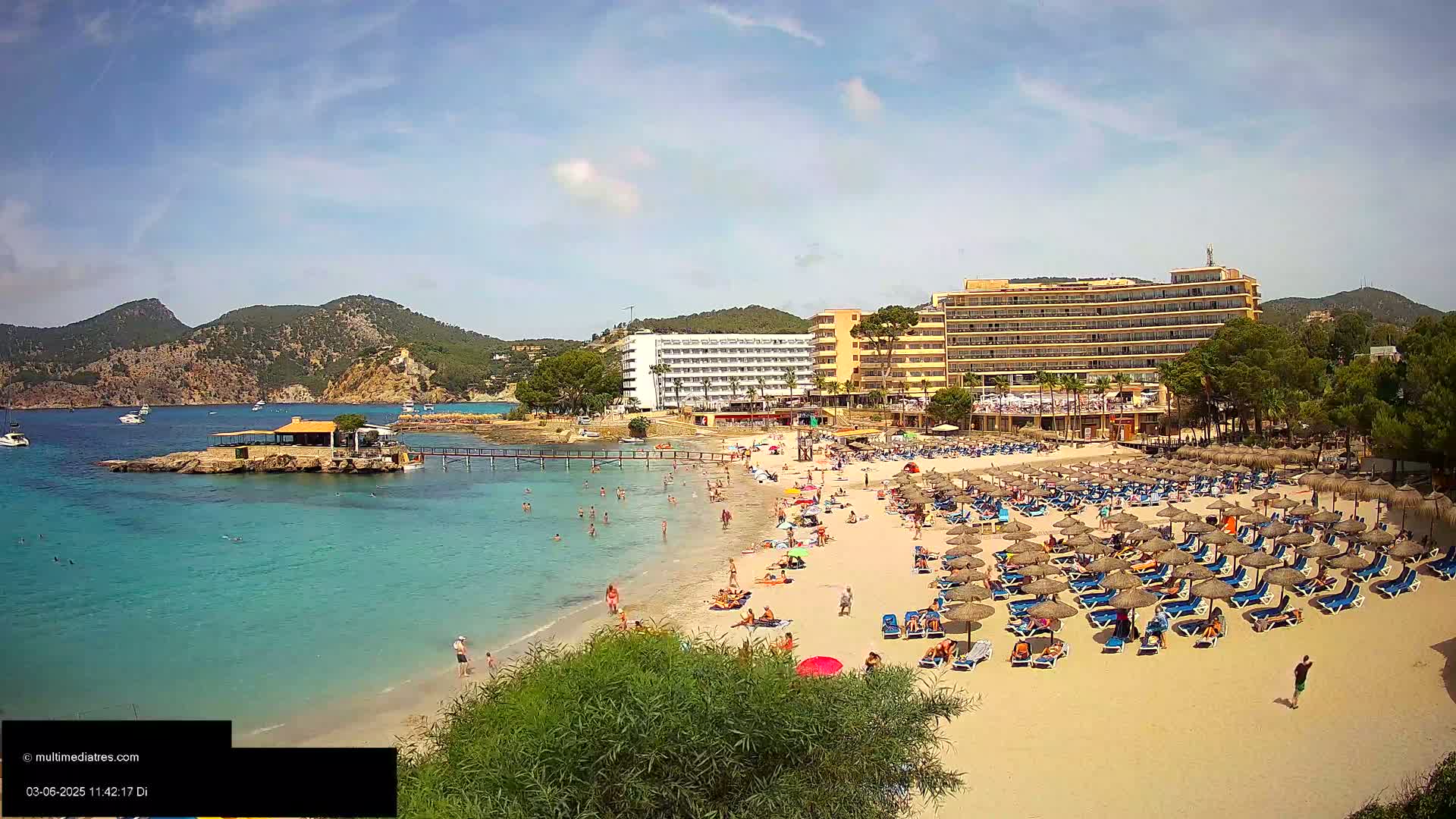 A sandy beach with numerous umbrellas and sunbathers is situated in front of a large hotel complex, all bordered by clear turquoise water and mountains under a mostly sunny sky.