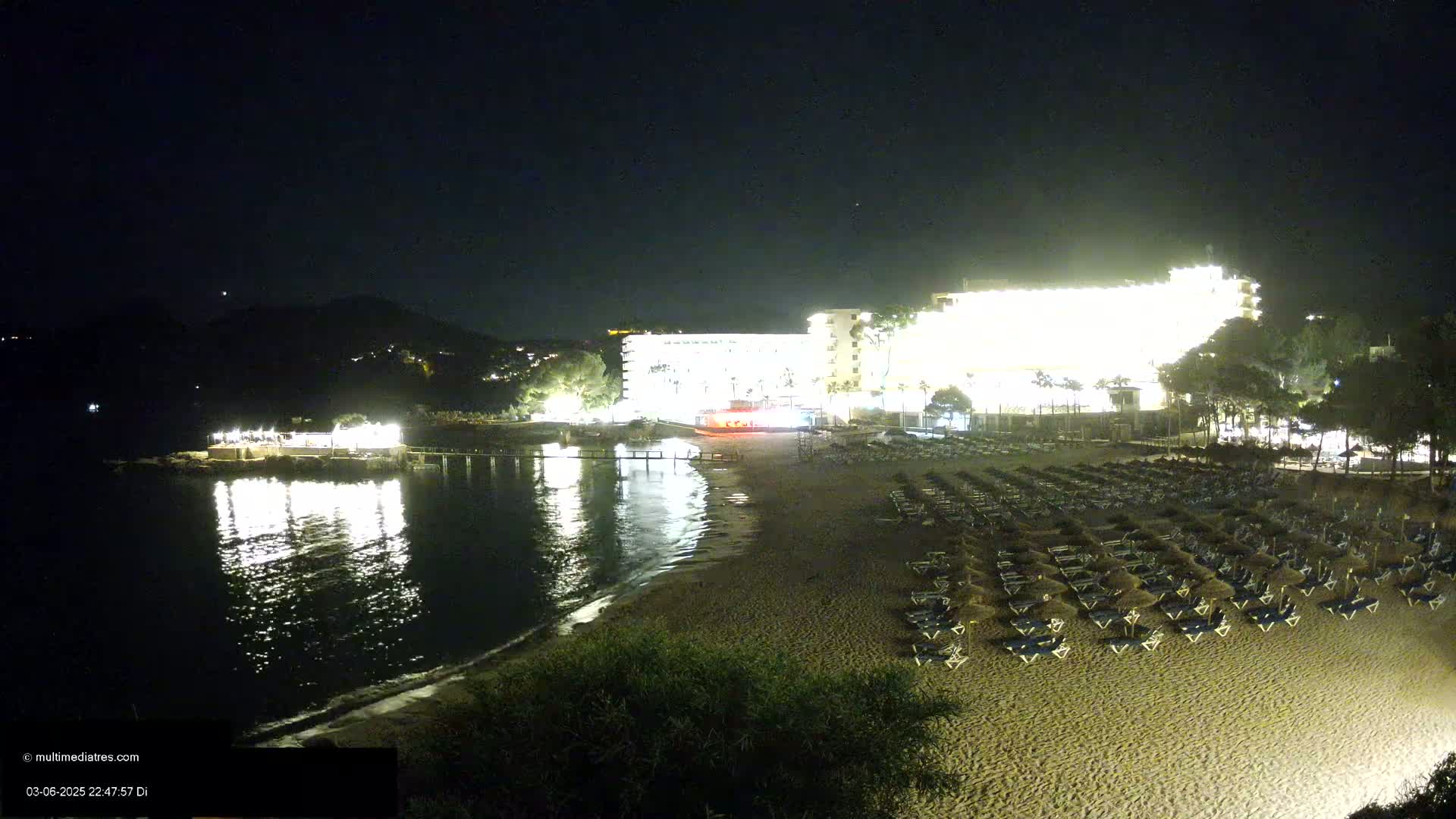 A brightly lit hotel overlooks a sandy beach at night, with calm water reflecting the lights.