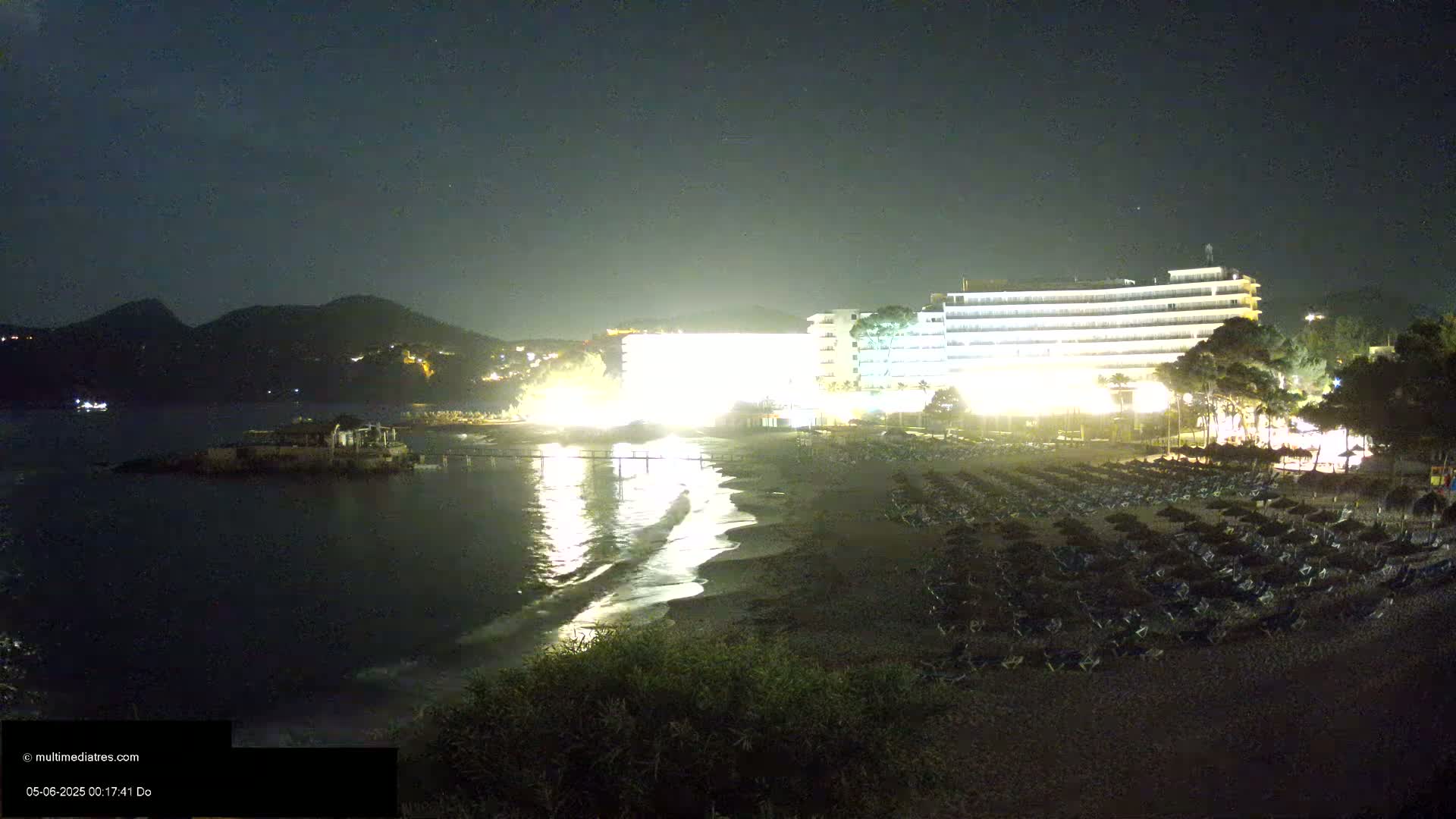 A brightly lit hotel overlooks a dark beach at night, with gently lapping waves on the shore and hills visible in the distance under a clear night sky.