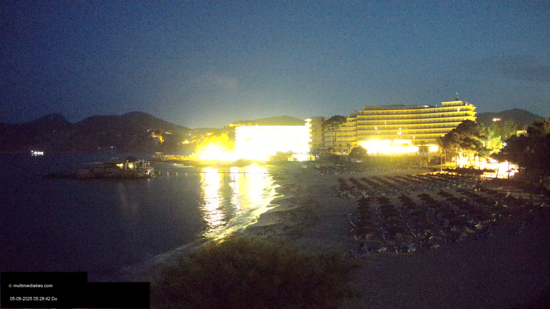 A brightly lit hotel and beach at night, with dark water and hills in the background under a clear, dark sky.