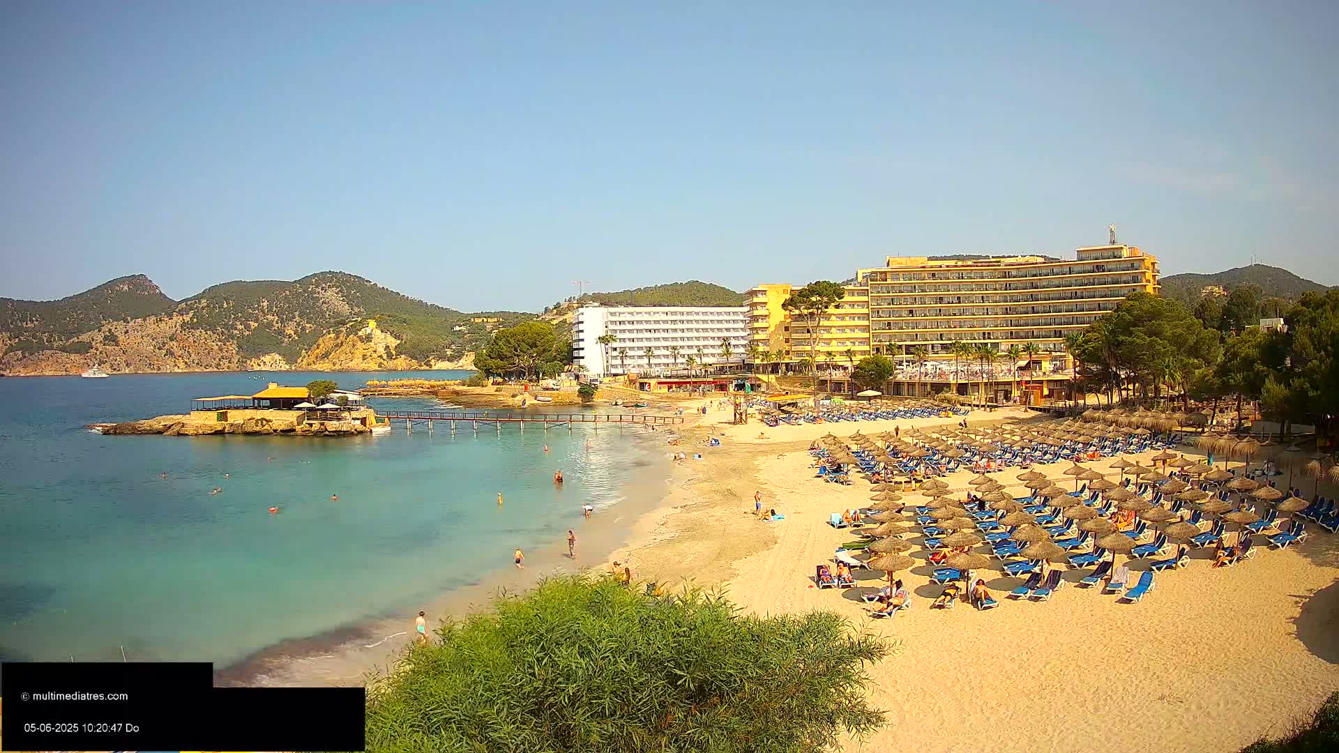 A sandy beach with numerous thatched umbrellas and lounge chairs is situated in front of several hotels, all under a clear, sunny sky, with calm, turquoise water and rocky outcroppings nearby.