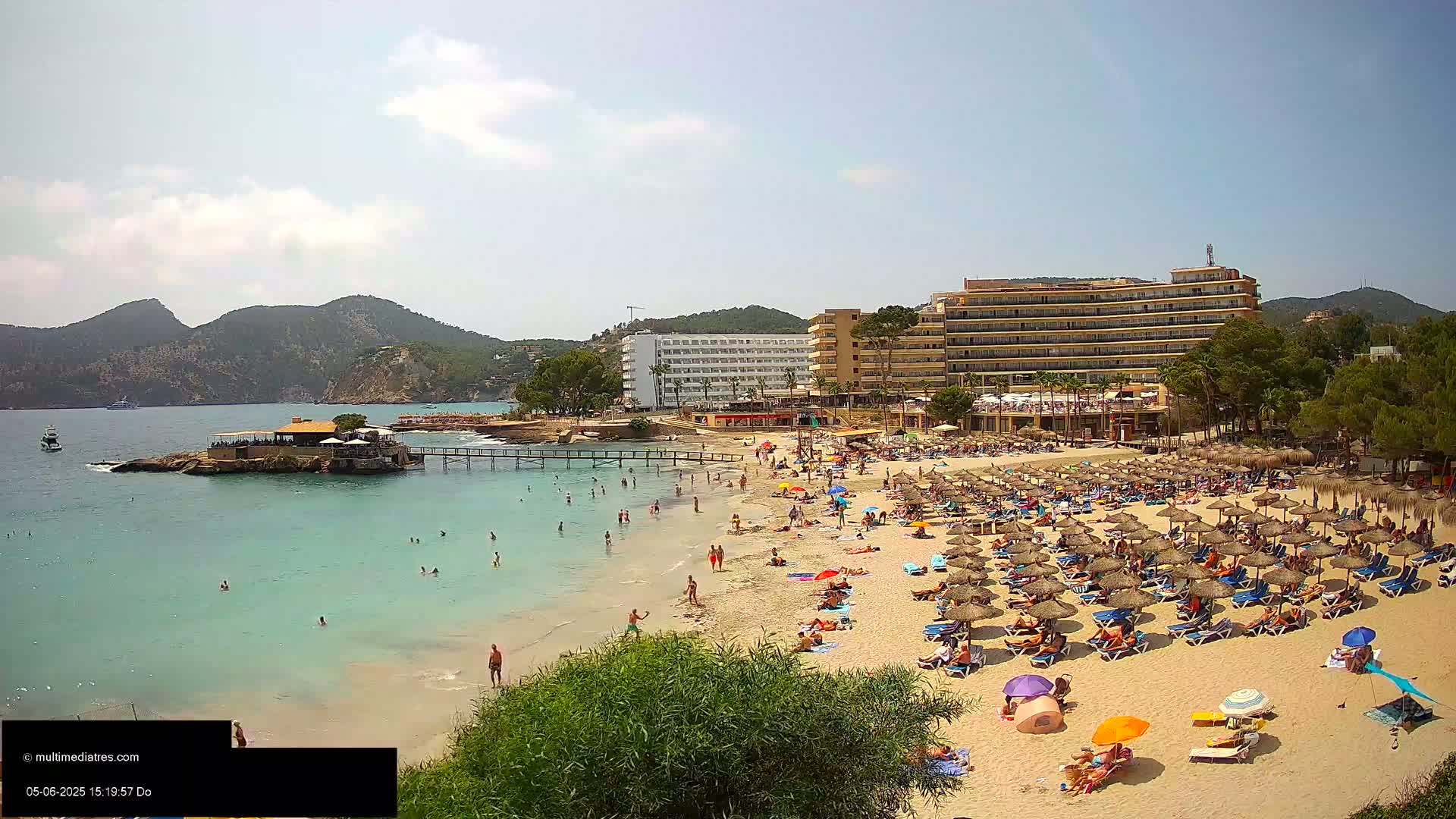 A sandy beach filled with sunbathers under numerous umbrellas and shade structures is situated next to a calm, turquoise body of water, backed by several hotels and lush green hills under a mostly sunny sky.