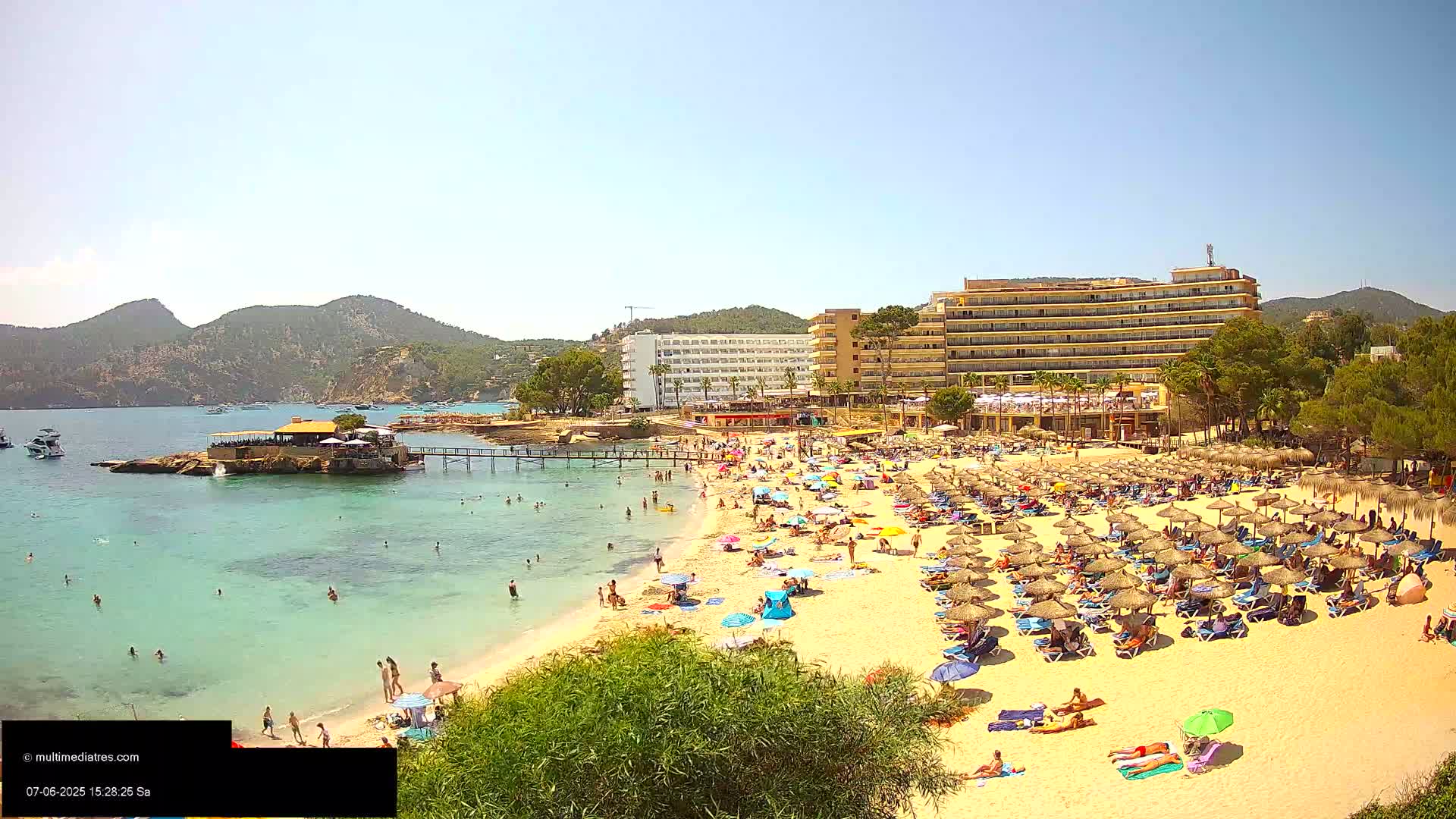 A sandy beach crowded with sunbathers under numerous umbrellas, bordered by turquoise water, a small island with a building, and large hotels, all under a clear sunny sky.
