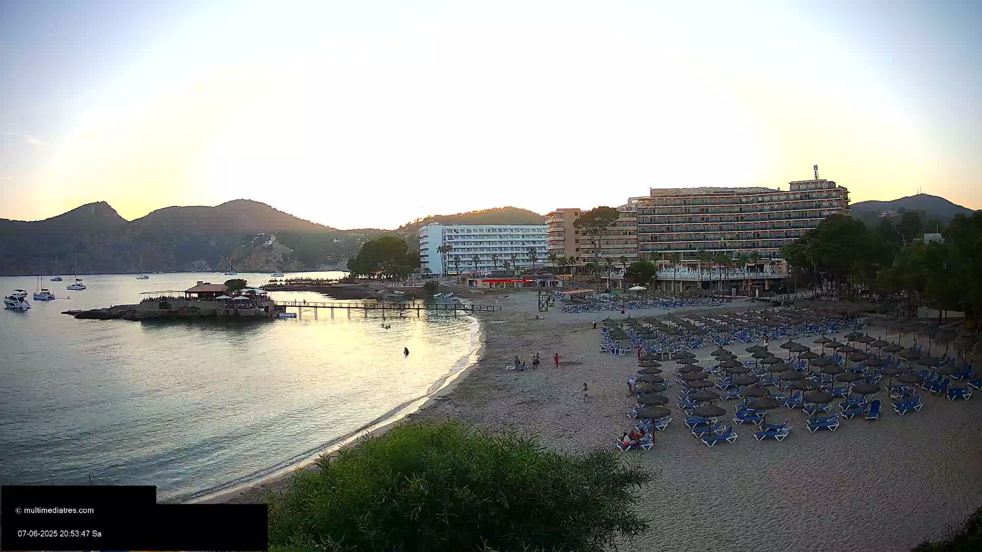 A sandy beach with numerous umbrellas and lounge chairs is situated in front of a large hotel, overlooking calm water and distant mountains under a clear twilight sky.