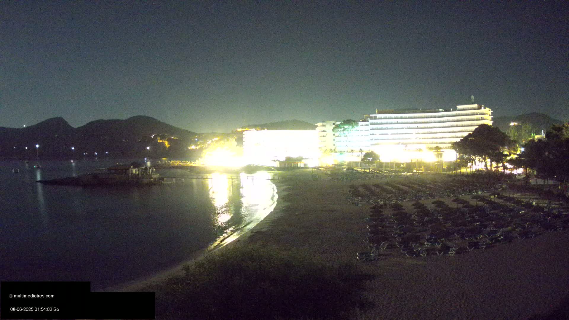 A brightly lit beachfront hotel at night overlooks a sandy beach strewn with lounge chairs, with dark hills and calm water visible in the background under a clear, dark sky.