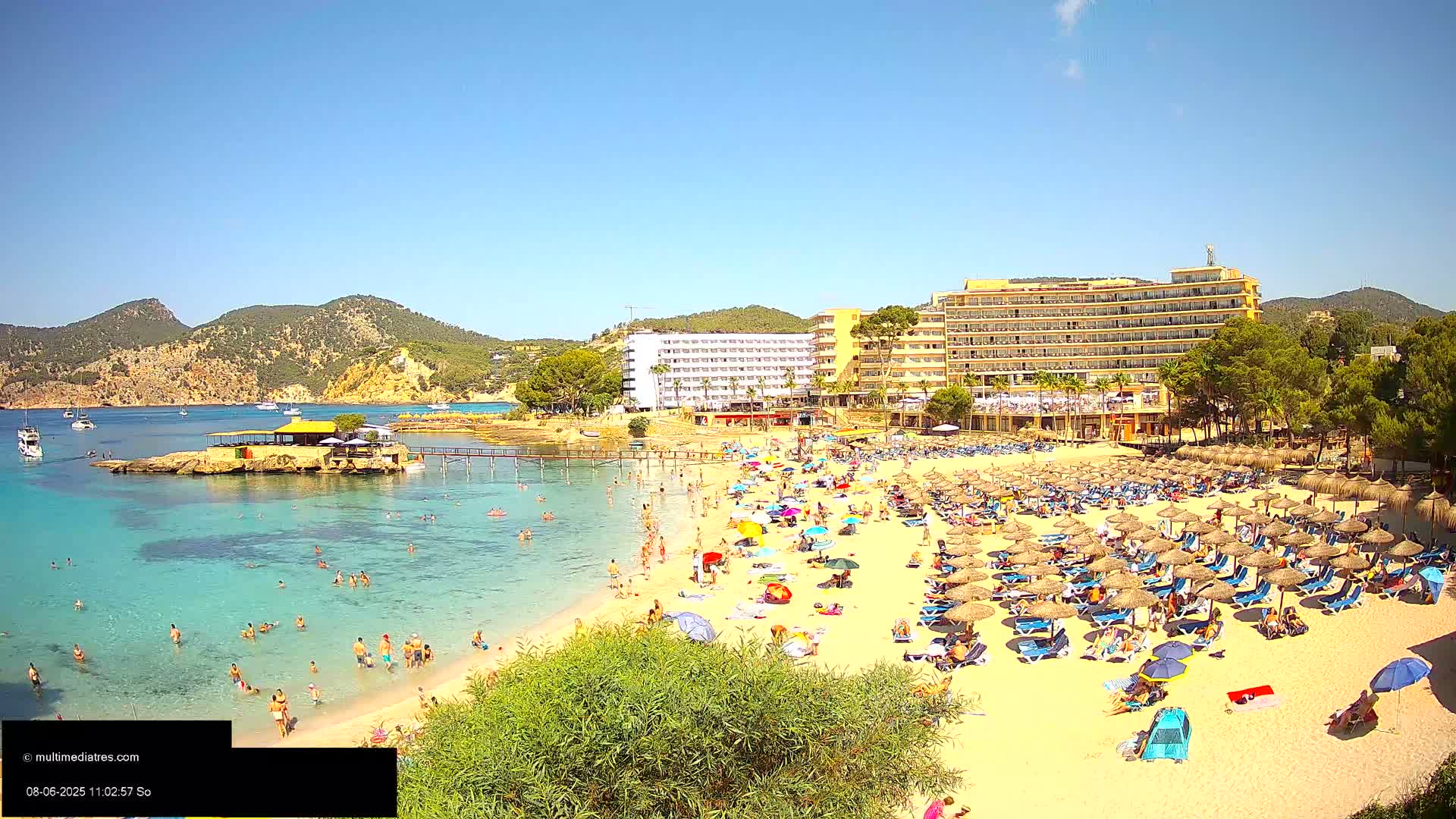 A crowded sandy beach with numerous thatched umbrellas and lounge chairs, clear turquoise water, and a large hotel behind it on a sunny day.