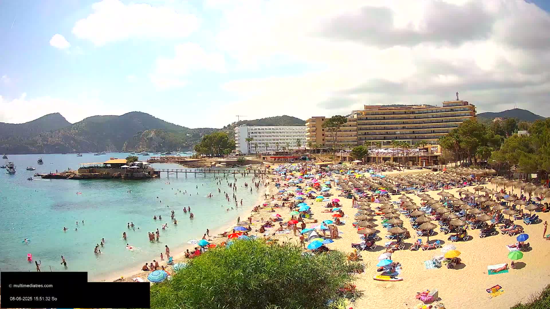 A crowded beach with numerous umbrellas and people swimming in clear, shallow water, backed by hotels and hills under a partly cloudy sky.