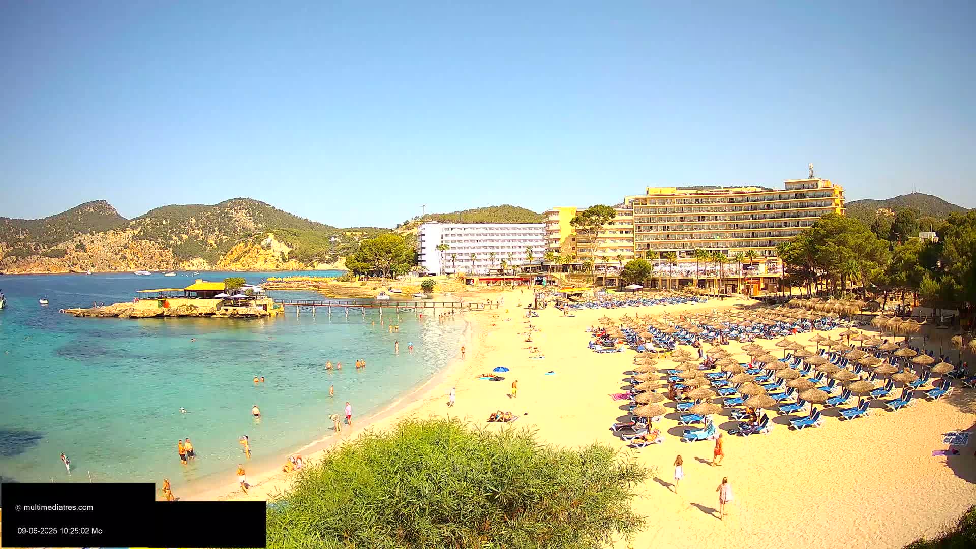 A sandy beach with many umbrellas and people, a calm turquoise ocean, and a large hotel situated on a hillside under a clear sunny sky.