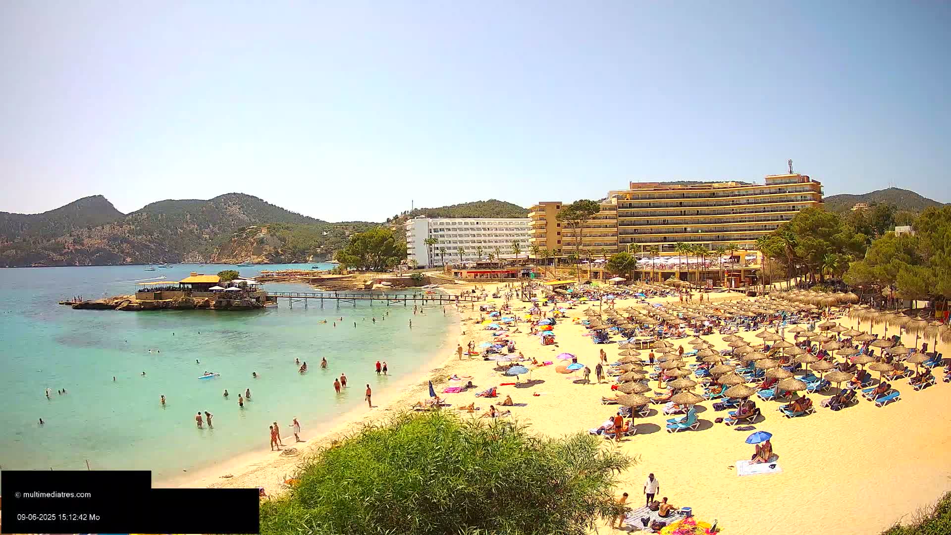 A sunny beach scene shows many people relaxing under umbrellas on a sandy beach, with turquoise water, a small island with a building, and several large hotels in the background.