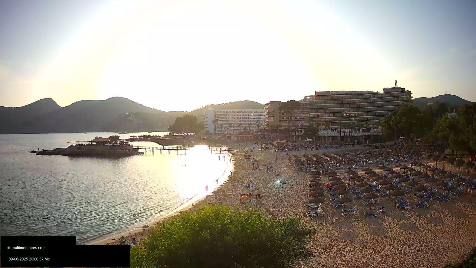 A sandy beach with numerous umbrellas and lounge chairs is situated in front of several hotels, with calm water and mountains in the background, under a mostly sunny sky.