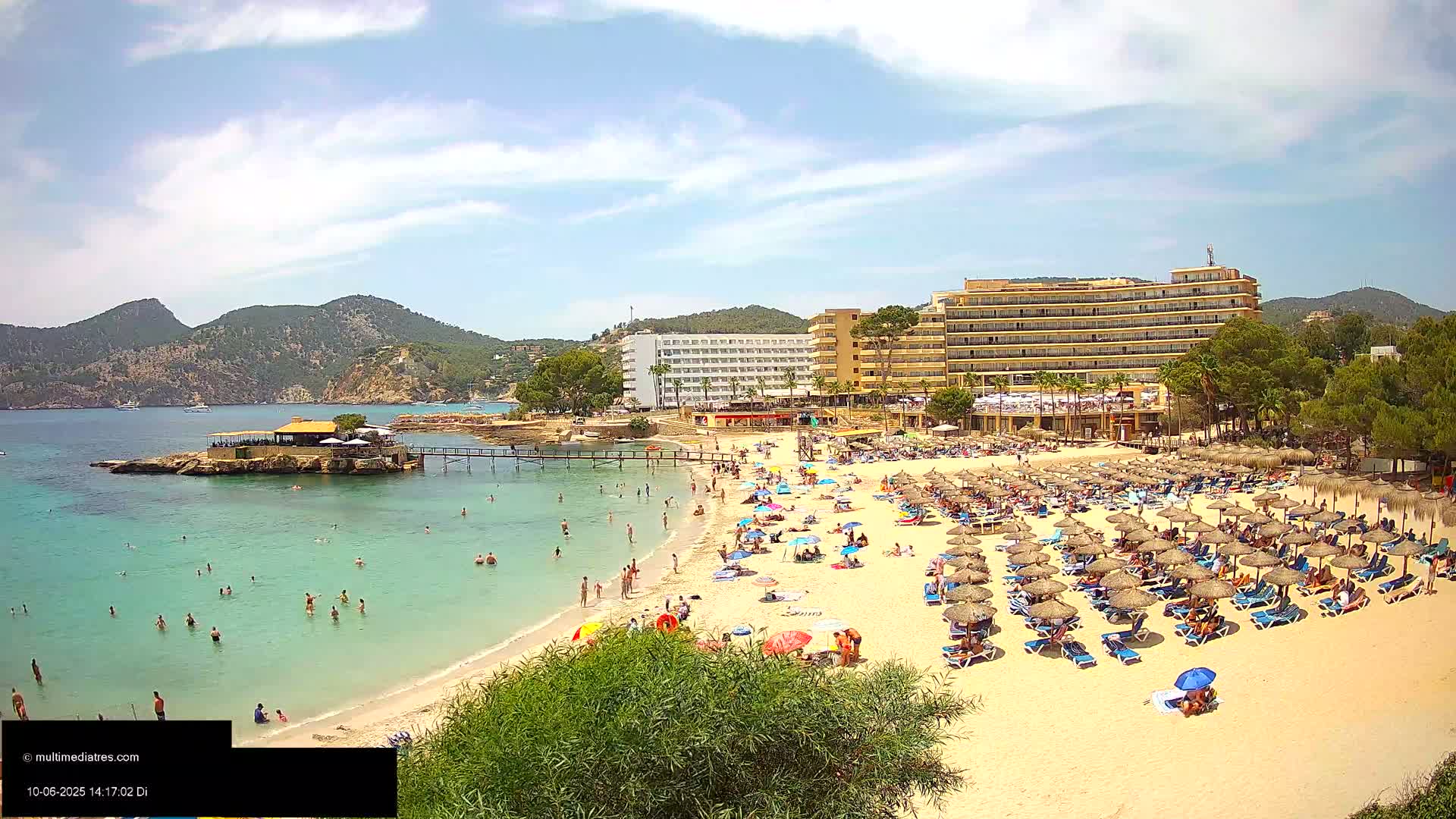A sandy beach with numerous umbrellas and sunbathers is situated near a large hotel and calm, clear turquoise water under a partly cloudy sky.