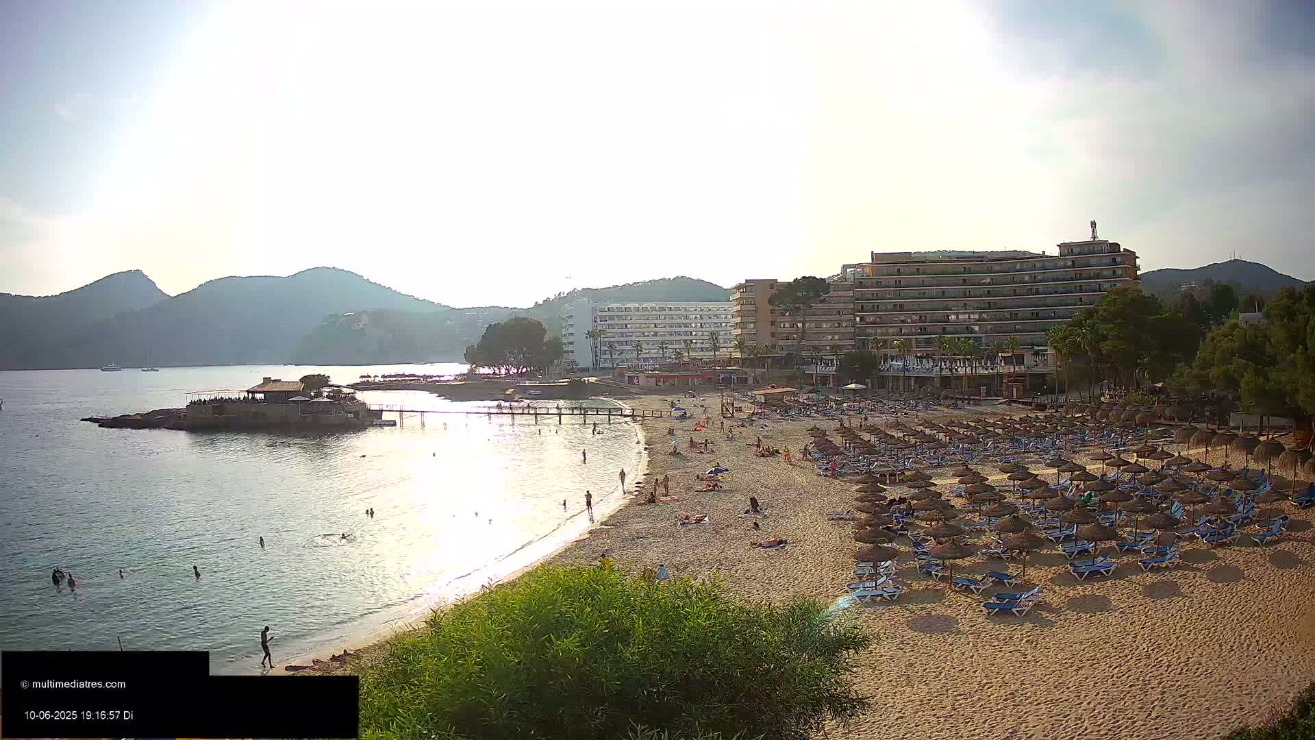 A sandy beach with numerous thatched umbrellas and lounge chairs is situated near calm, clear water, backed by hotels and hills under a bright, sunny sky.