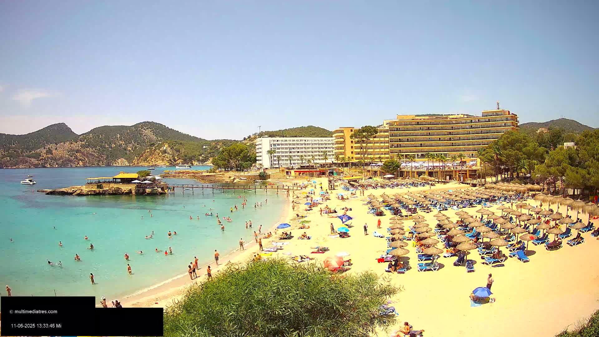 A sunny beach scene shows a sandy shore crowded with beachgoers under numerous umbrellas, a calm turquoise ocean with swimmers, and hotels and vegetation in the background.