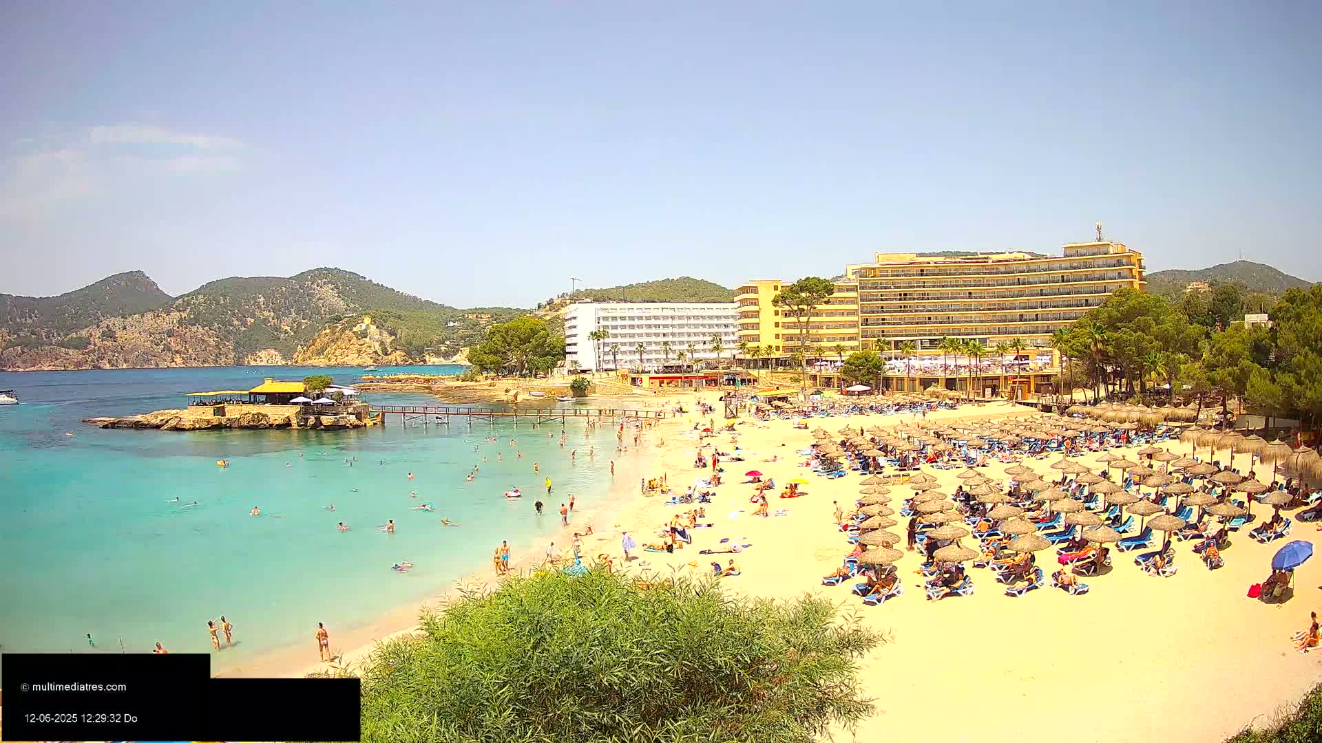A sunny beach scene shows numerous people relaxing under beach umbrellas near a resort, with calm, clear turquoise water and rocky hills in the background.
