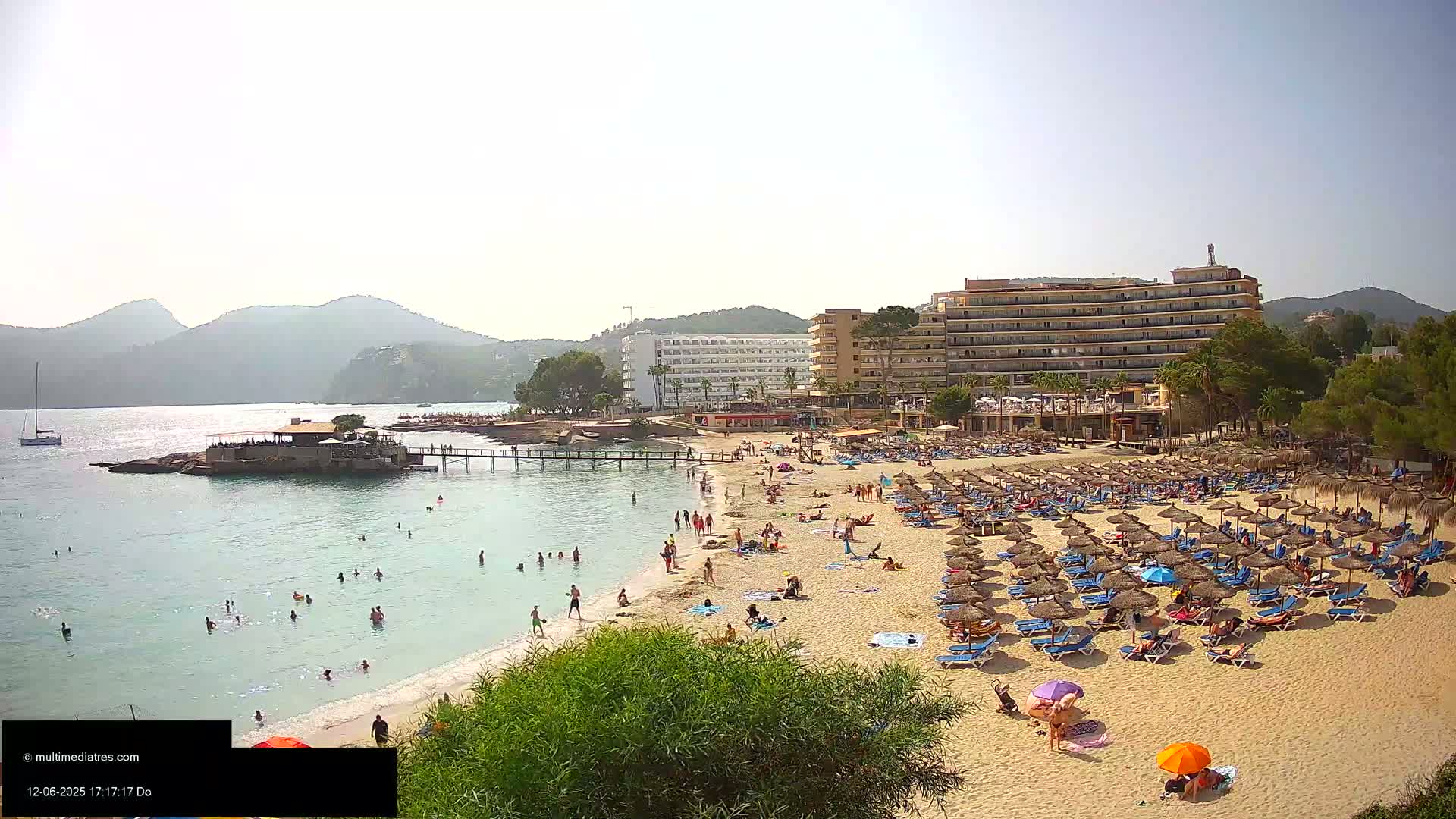 A sandy beach with many people, umbrellas, and lounge chairs is situated in front of several hotels and a calm, clear body of water, backed by hills under a sunny sky.