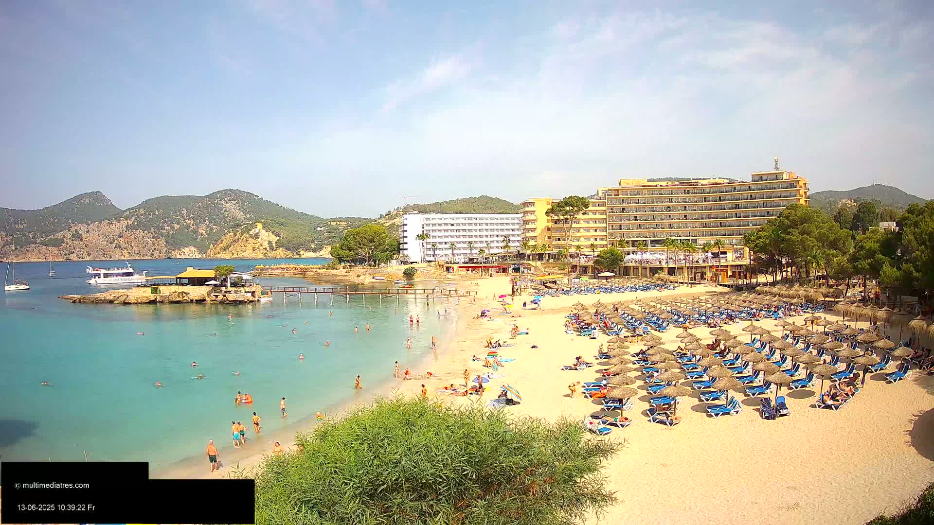 A sandy beach with numerous thatched umbrellas and lounge chairs, a large hotel, and people swimming and sunbathing in clear turquoise water under a bright sunny sky, backed by hills.