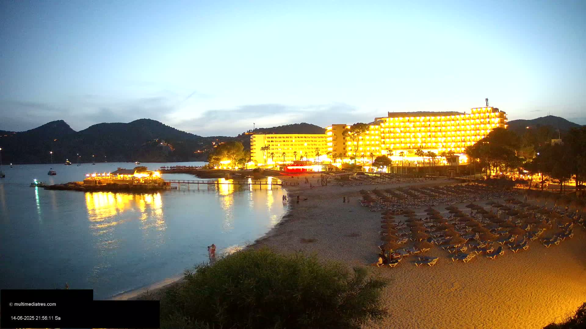 A sandy beach with numerous thatched umbrellas and lounge chairs, a large hotel, and people swimming and sunbathing in clear turquoise water under a bright sunny sky, backed by hills.