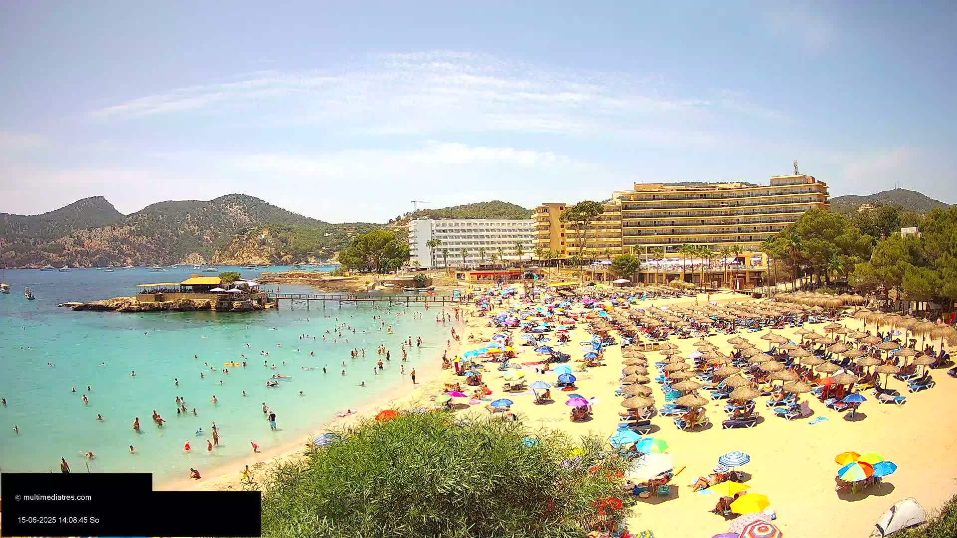 A crowded beach with turquoise water, many umbrellas and beach chairs, and hotels behind it on a sunny day.