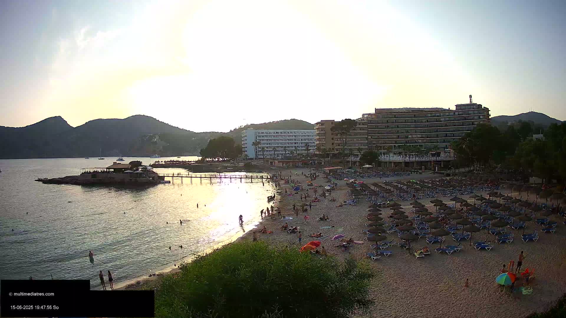 A sandy beach with numerous lounge chairs and umbrellas under a setting sun, adjacent to a calm bay with a small island featuring a building and a pier, backed by several hotels and hills.