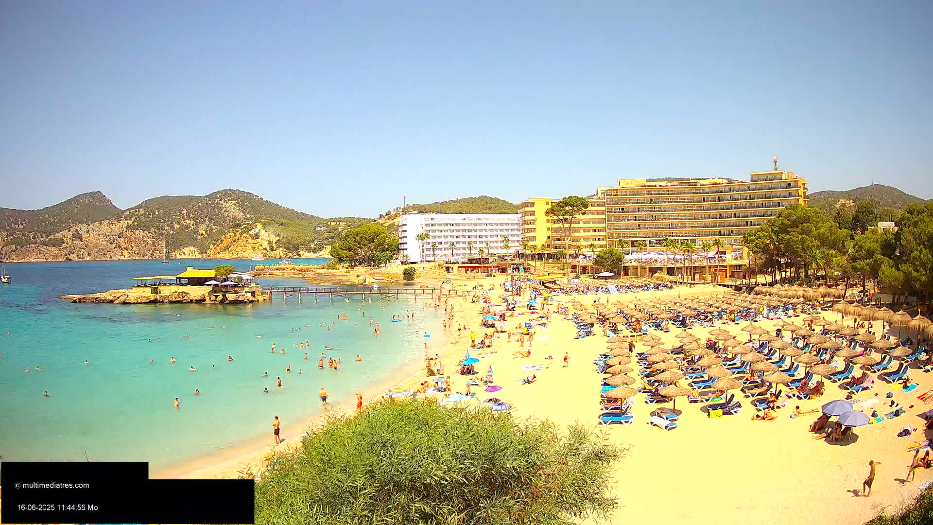 A sunny beach scene shows a sandy shore crowded with beachgoers under numerous thatched umbrellas, a calm turquoise ocean with swimmers, and hotels and vegetation behind the beach.
